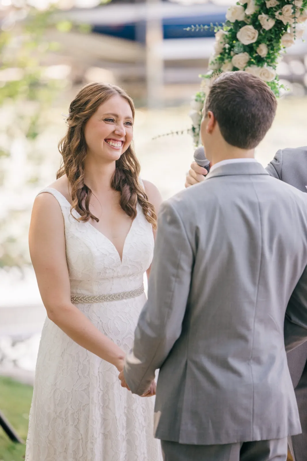 The bride beams at her groom during the ceremony as he speaks into a microphone beneath a white-rose arch, with a lake and dock blurred behind them. — Tim Larsen Photography, Brainerd Lakes MN