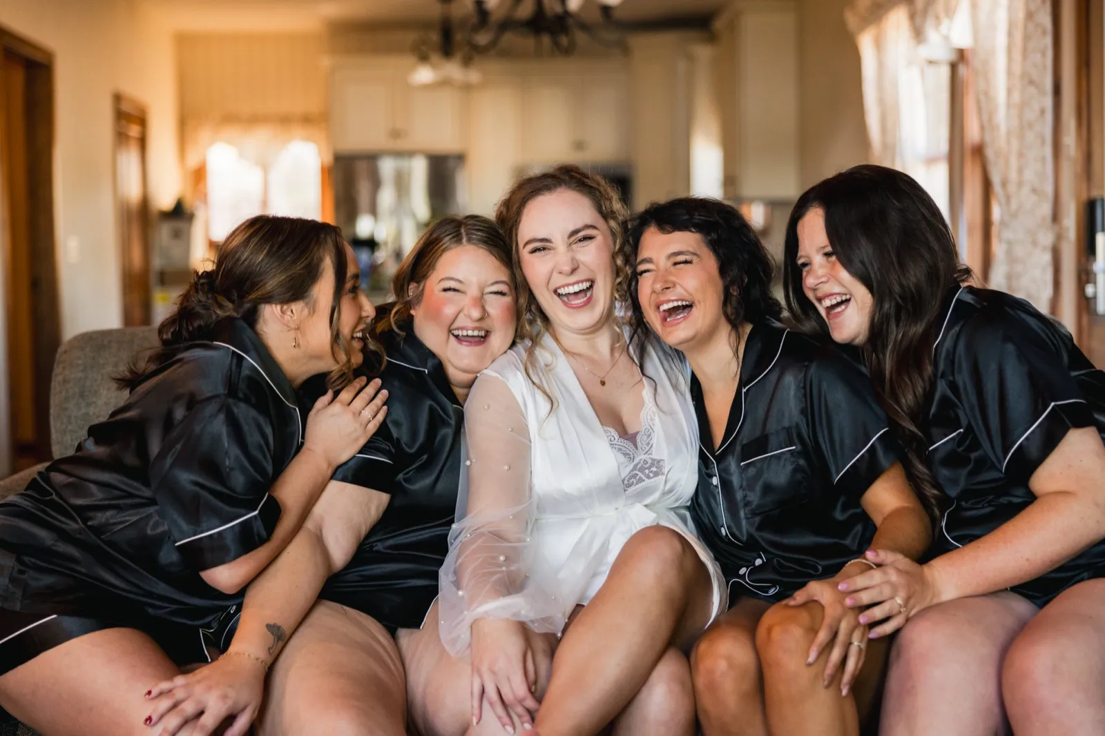 The bride sits on a couch in a sheer white robe surrounded by four bridesmaids in black satin pajamas, all laughing together in window light — Tim Larsen Photography, Brainerd Lakes MN