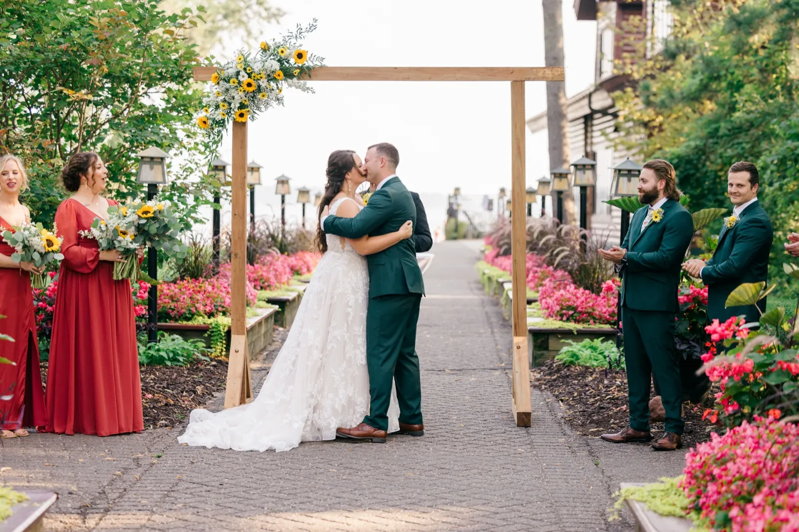 Chelsea and Eric kiss beneath a sunflower-draped wooden arch as their bridal party watches and reacts on a stone garden path at Grand View Lodge — Tim Larsen Photography, Brainerd Lakes MN