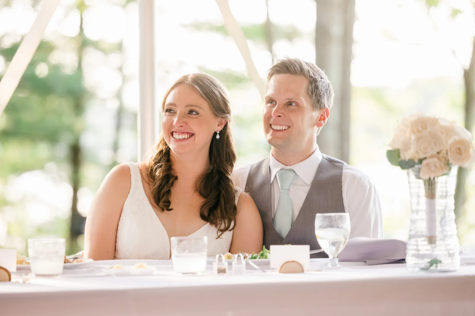 The bride and groom laugh together at their sweetheart table during toasts, a white rose bouquet visible to their right. — Tim Larsen Photography, Brainerd Lakes MN