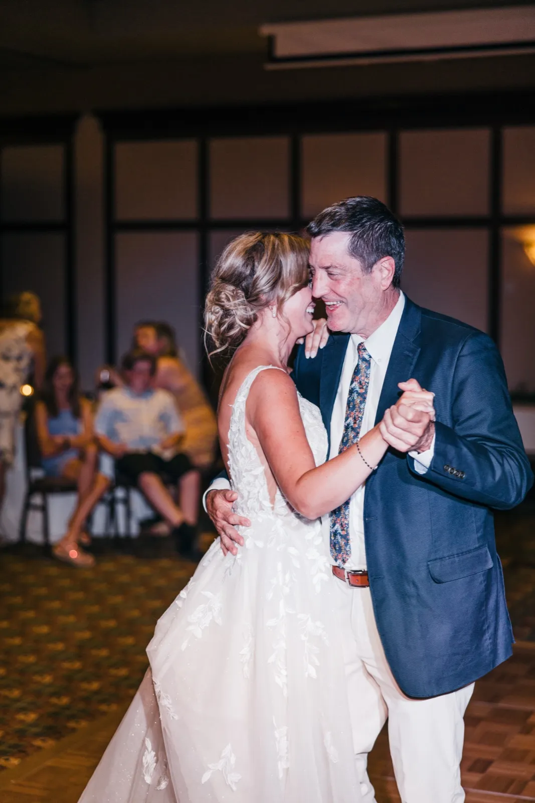 The bride and her father laugh forehead-to-forehead while dancing together on the reception floor as seated guests watch from behind.