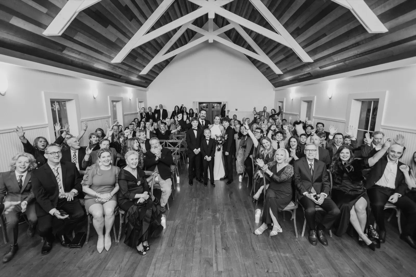 The couple stands at the center of every wedding guest gathered in a vaulted-ceiling hall, the whole room turned toward the camera with arms raised — the all-in group frame, caught before anyone scatters — Tim Larsen Photography, Brainerd Lakes MN