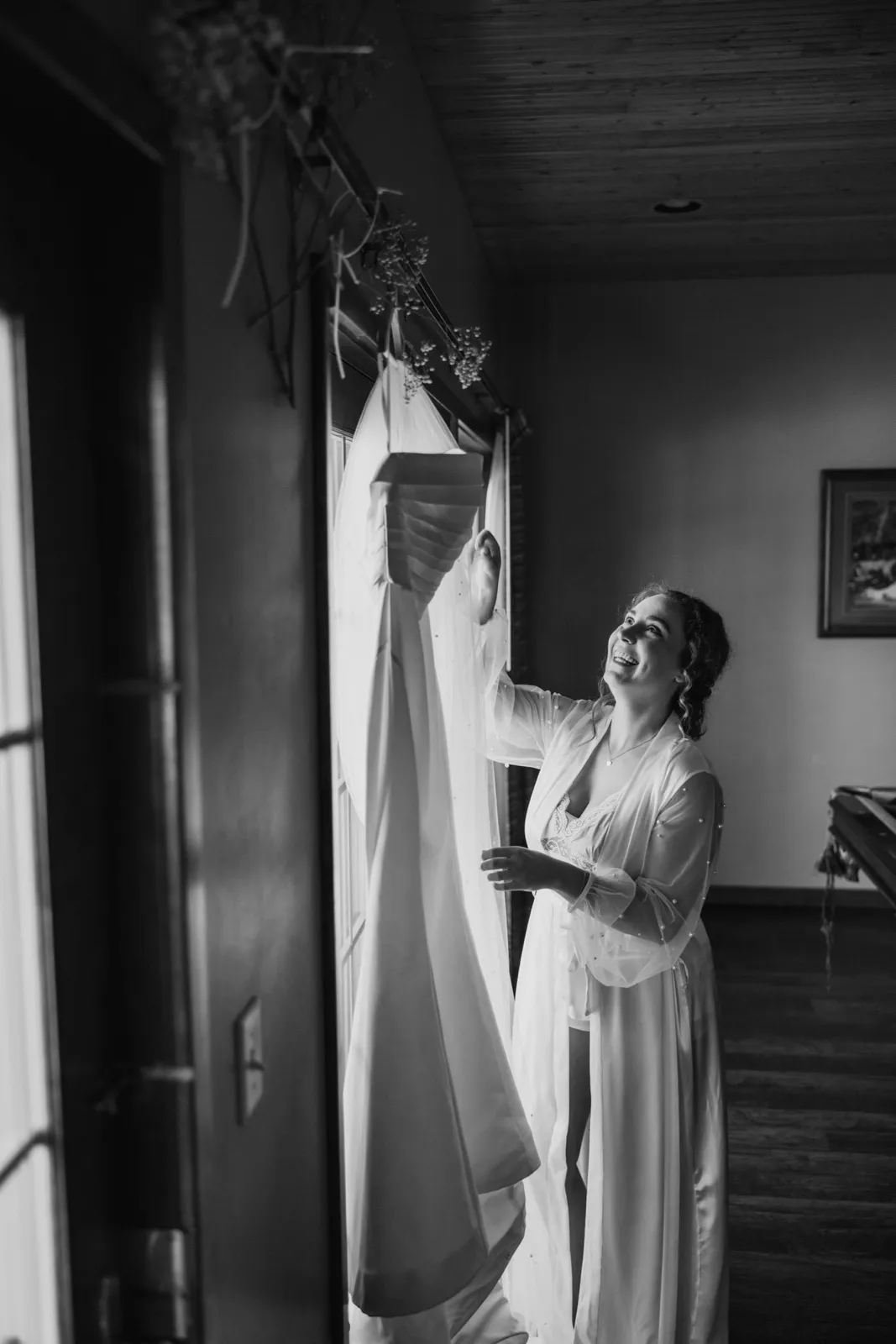The bride in a silk robe reaches up toward her wedding dress hanging in a window, smiling and looking up at it — Tim Larsen Photography, Brainerd Lakes MN