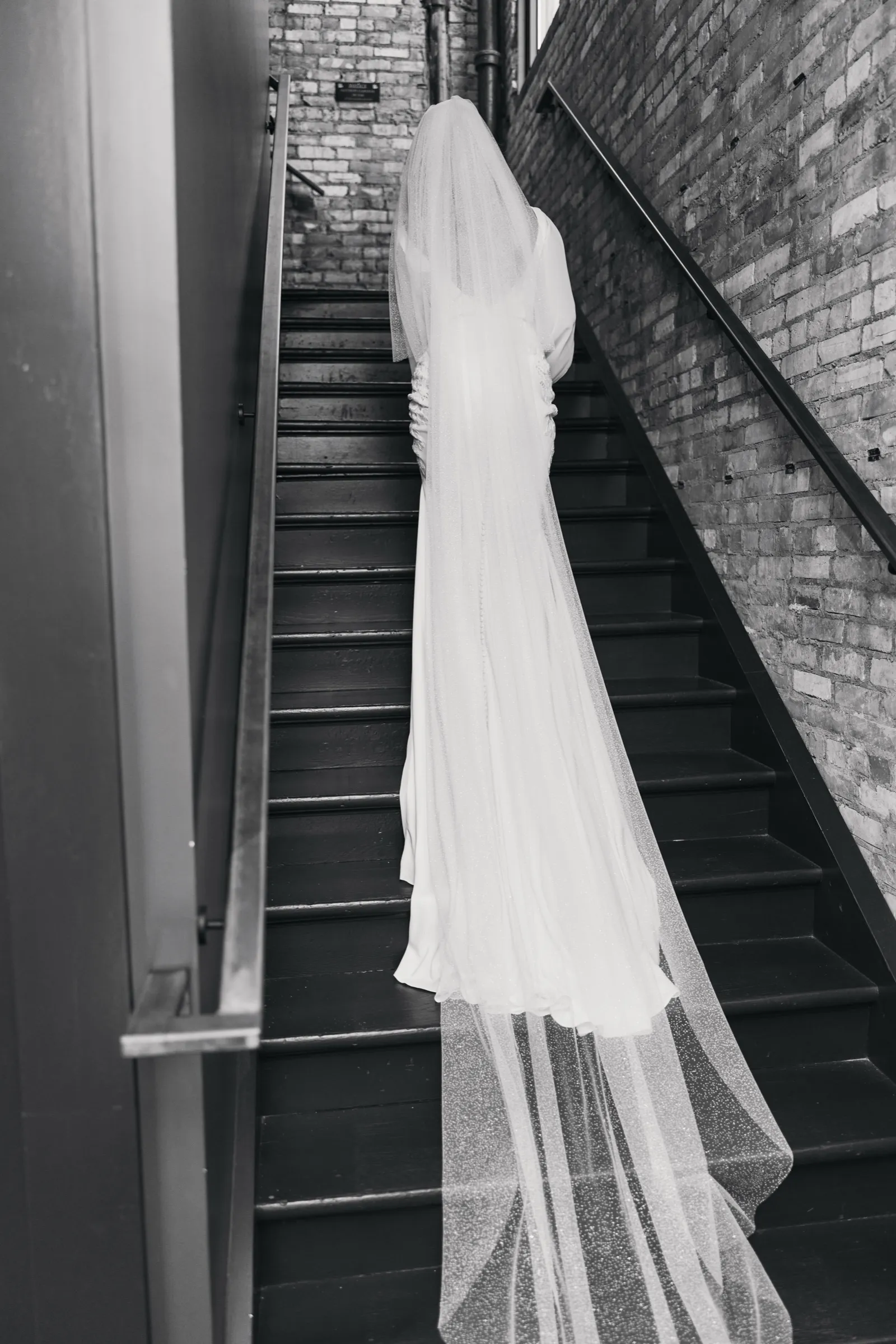 Madeline climbs a dark wooden staircase in a brick stairwell, her long veil and gown trailing down the steps behind her — Tim Larsen Photography, Brainerd Lakes MN
