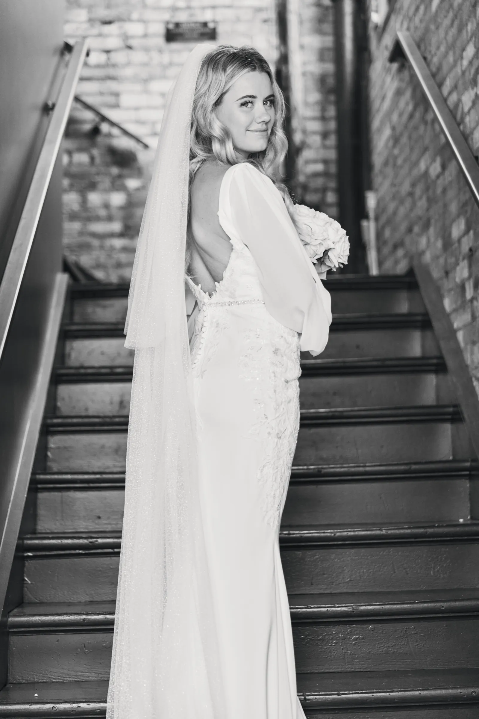 Madeline looks back over her shoulder on a brick stairway, her long veil trailing down the steps behind her — Tim Larsen Photography, Brainerd Lakes MN