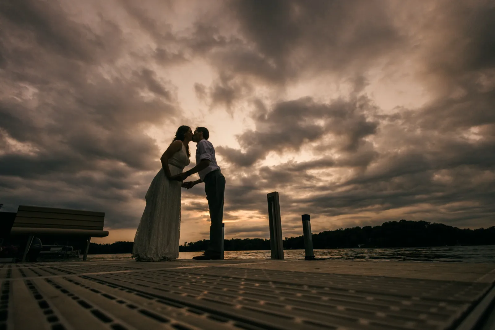 The couple kisses in silhouette on a wooden dock beneath a vast, dramatic cloudy sky over a lake at dusk. — Tim Larsen Photography, Brainerd Lakes MN