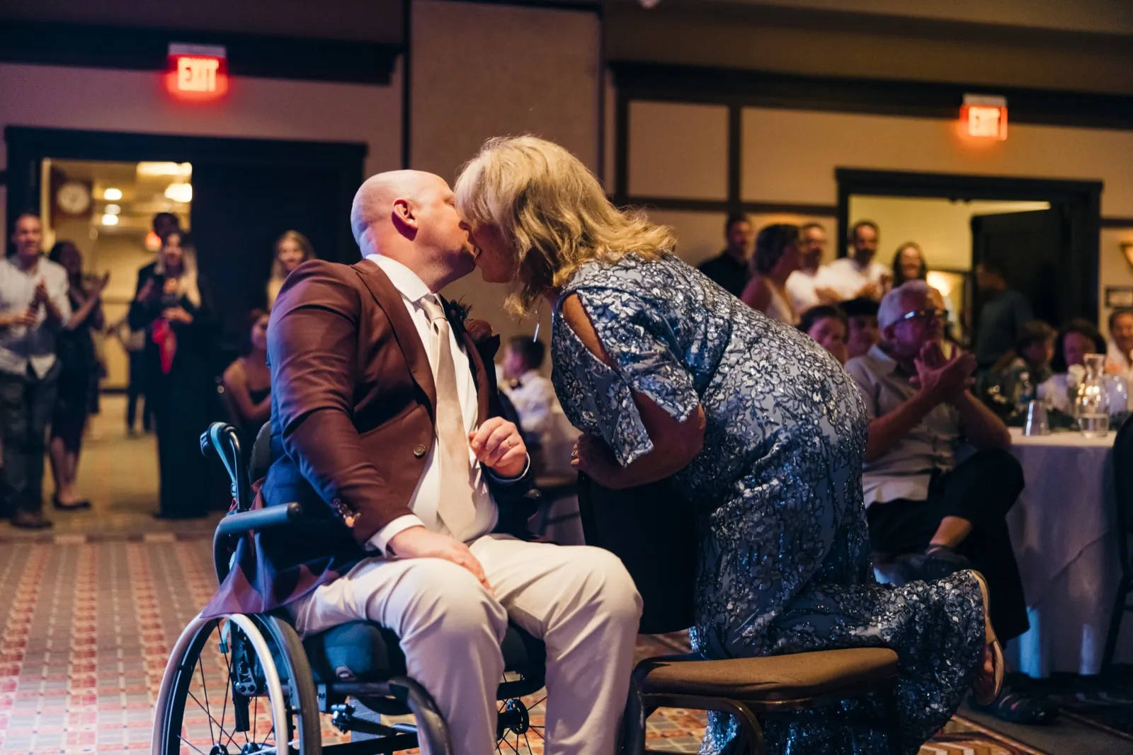 The groom, seated in a wheelchair, leans up to kiss his mother as guests watch from tables and the doorway during the reception. — Tim Larsen Photography, Brainerd Lakes MN