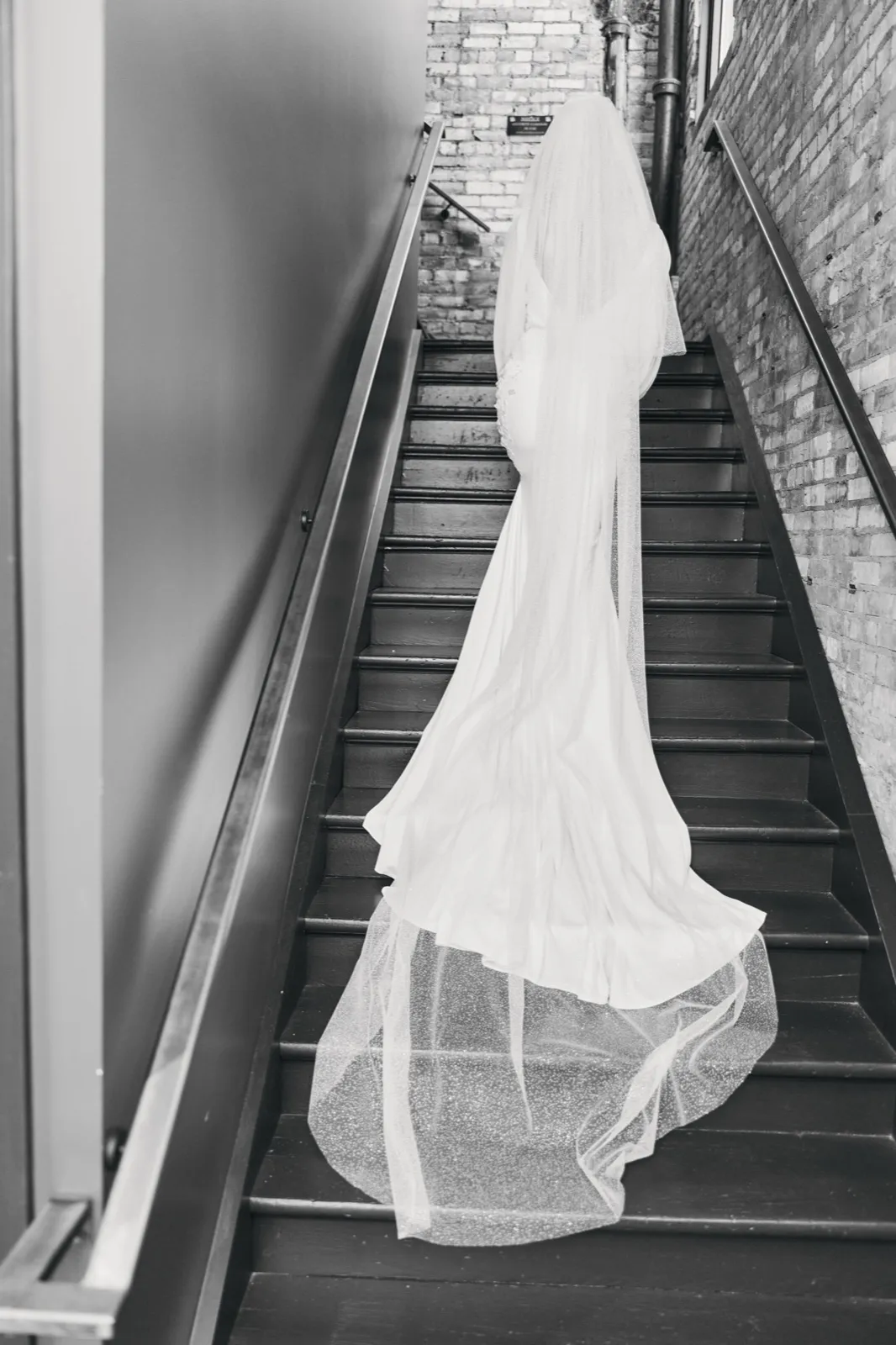 The bride ascends a wooden staircase in a brick-walled stairwell, her long veil and train cascading down the steps behind her, photographed in black and white. — Tim Larsen Photography, Brainerd Lakes MN