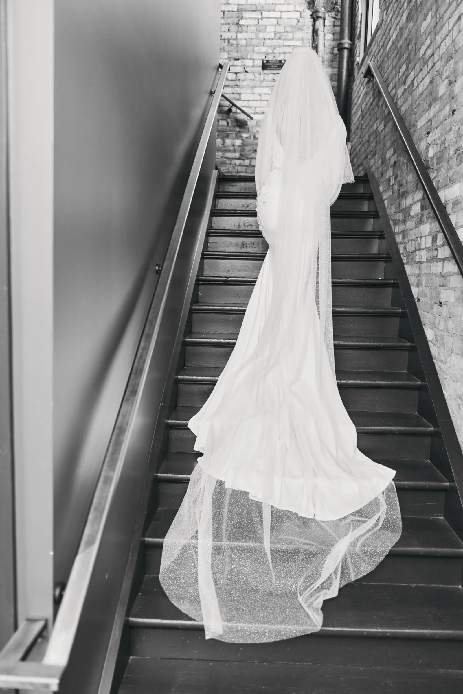 Madeline ascends a wooden staircase in a brick-walled stairwell in black and white, her long veil and train cascading down the steps behind her — Tim Larsen Photography, Brainerd Lakes MN