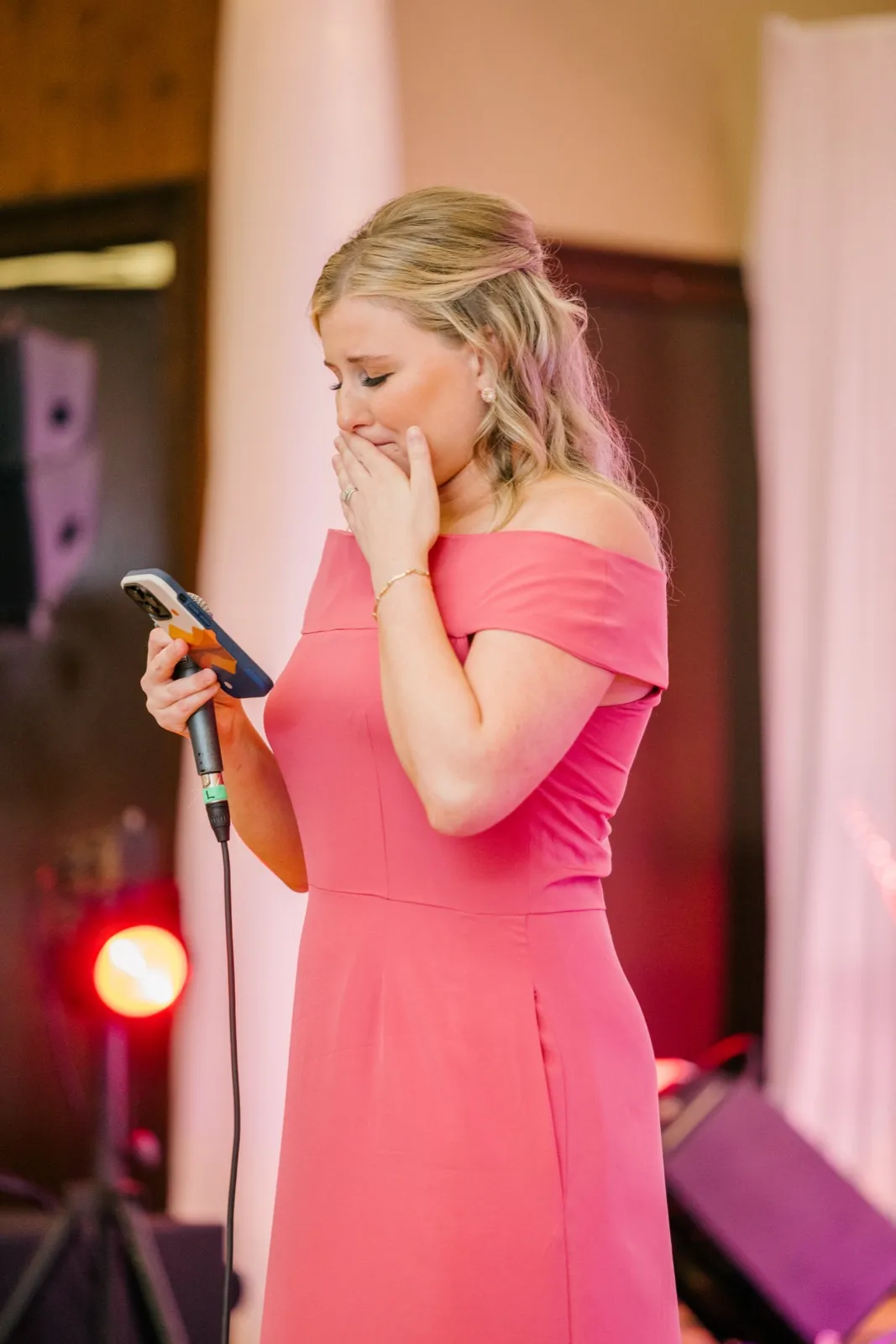 A bridesmaid in a pink off-shoulder dress covers her mouth, overcome with emotion mid-toast as she holds a phone and microphone — Tim Larsen Photography, Brainerd Lakes MN