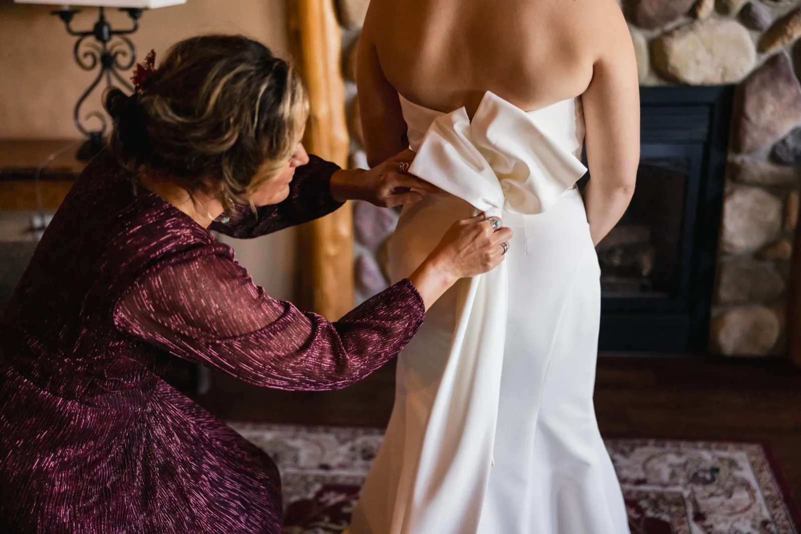 A woman in a burgundy dress kneels to fasten the back of the bride's white gown in a room with a stone fireplace — Tim Larsen Photography, Brainerd Lakes MN