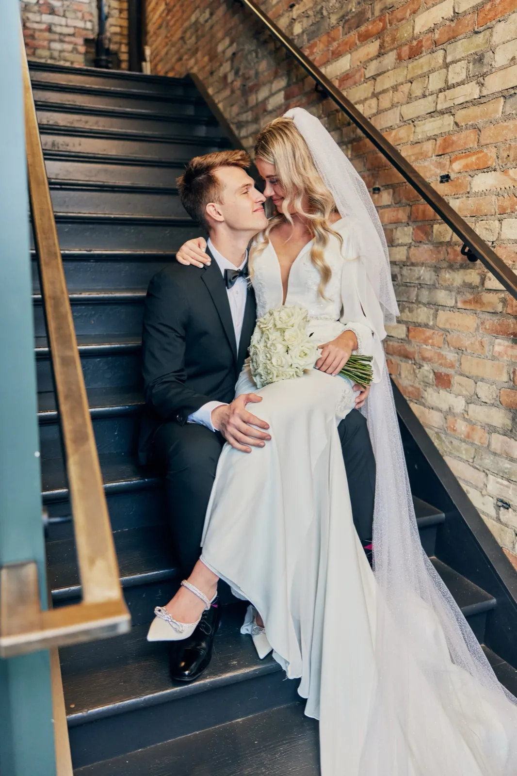The bride and groom sit close together on a dark wooden staircase against an exposed brick wall, foreheads touching, with her veil and gown cascading down the steps. — Tim Larsen Photography, Brainerd Lakes MN