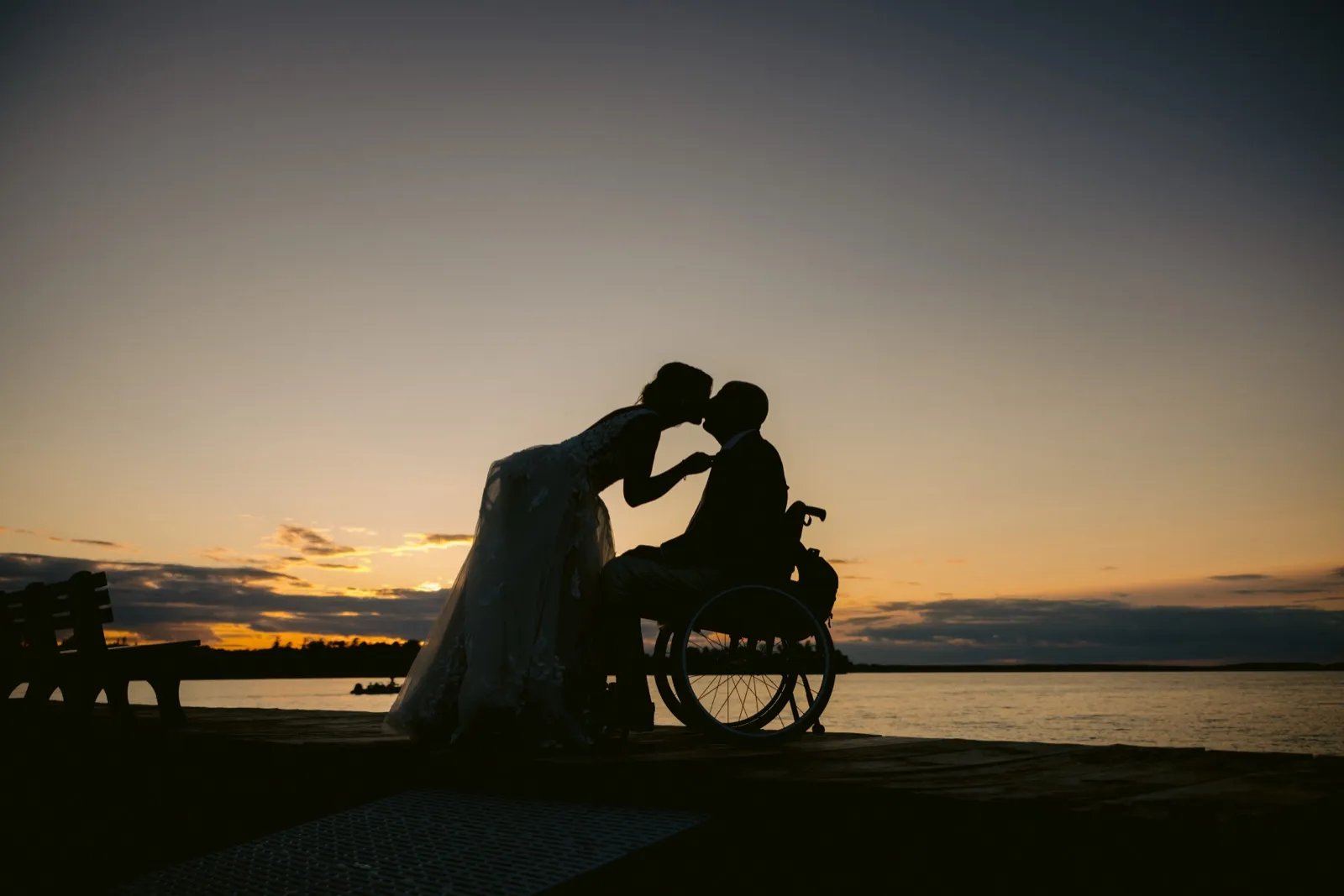 The bride leans down to kiss the groom, who is seated in a wheelchair on a lakeside dock, both silhouetted against a dusk sky over the water. — Tim Larsen Photography, Brainerd Lakes MN
