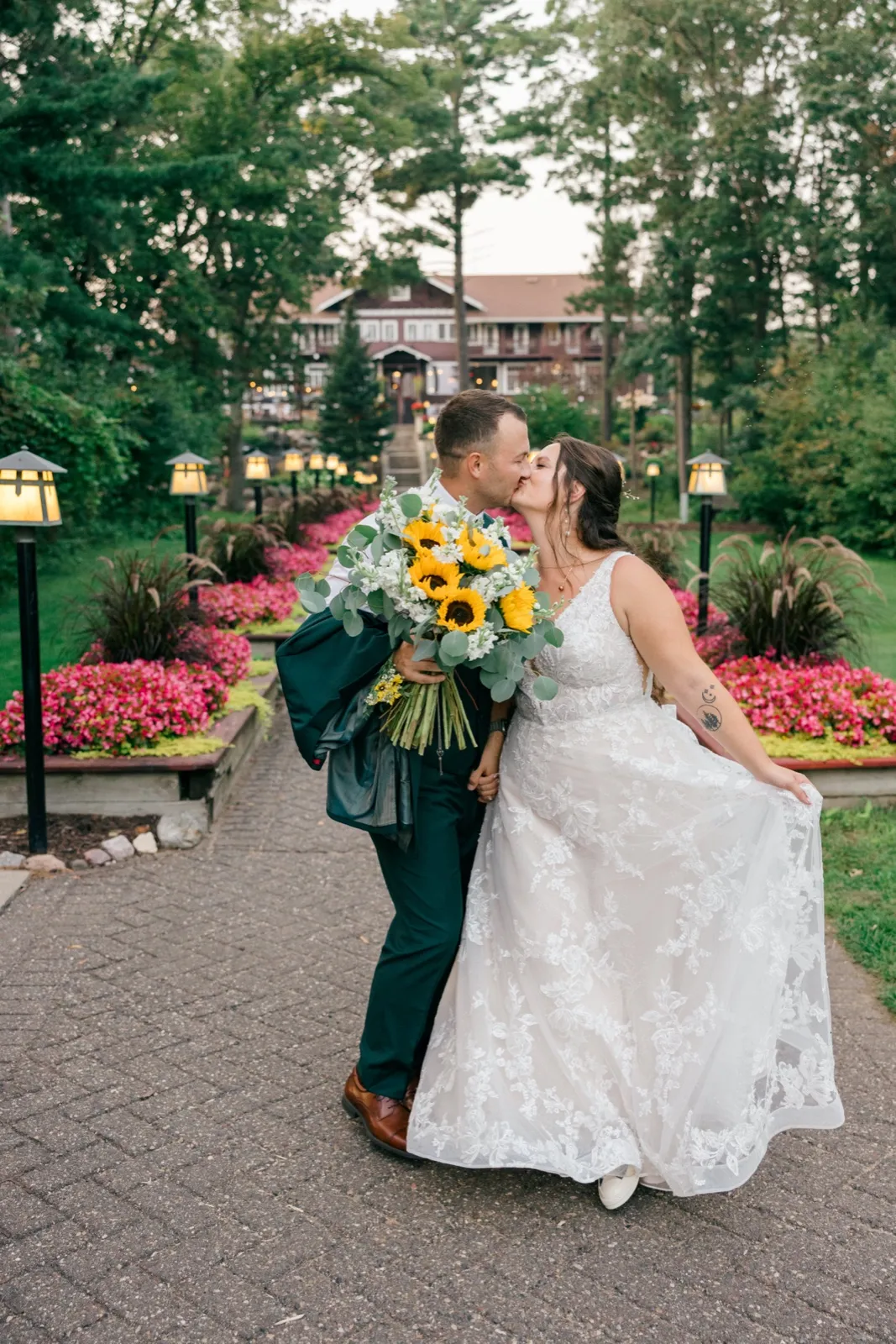 Chelsea and Eric share a kiss on a lantern-lined brick pathway with the historic Grand View Lodge main building visible in the background, the bride holding a large sunflower bouquet — Tim Larsen Photography, Brainerd Lakes MN