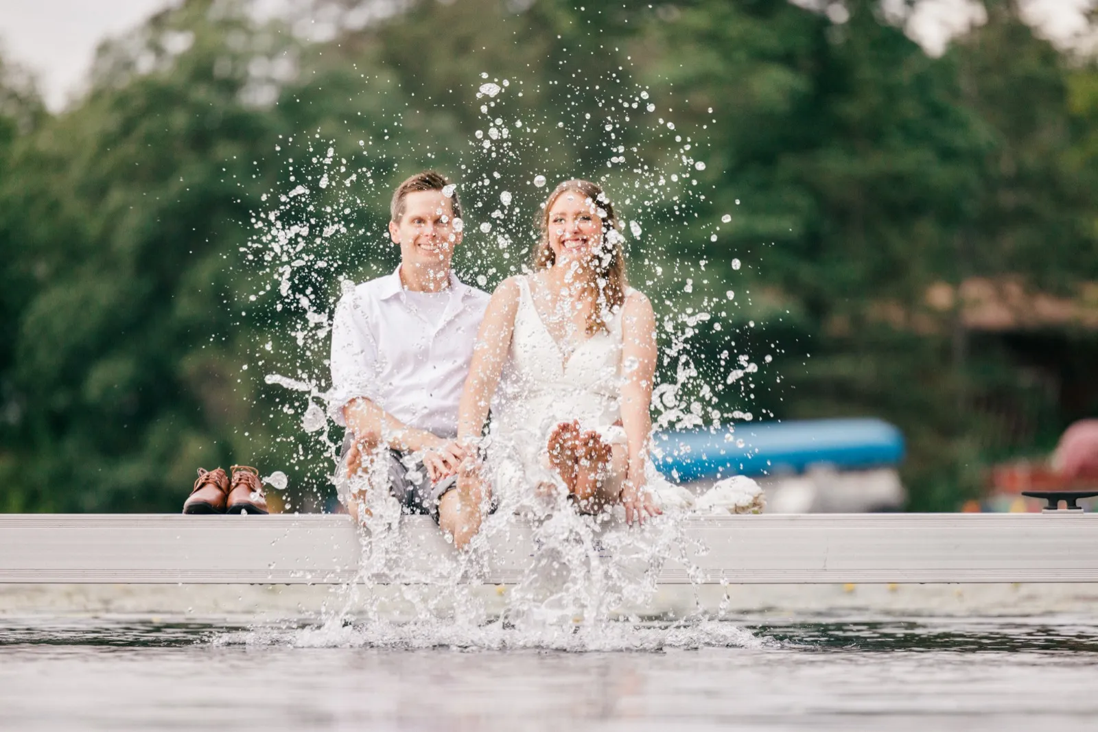 The bride and groom sit at the edge of a dock kicking up a large splash of lake water with their bare feet, both laughing toward the camera. — Tim Larsen Photography, Brainerd Lakes MN