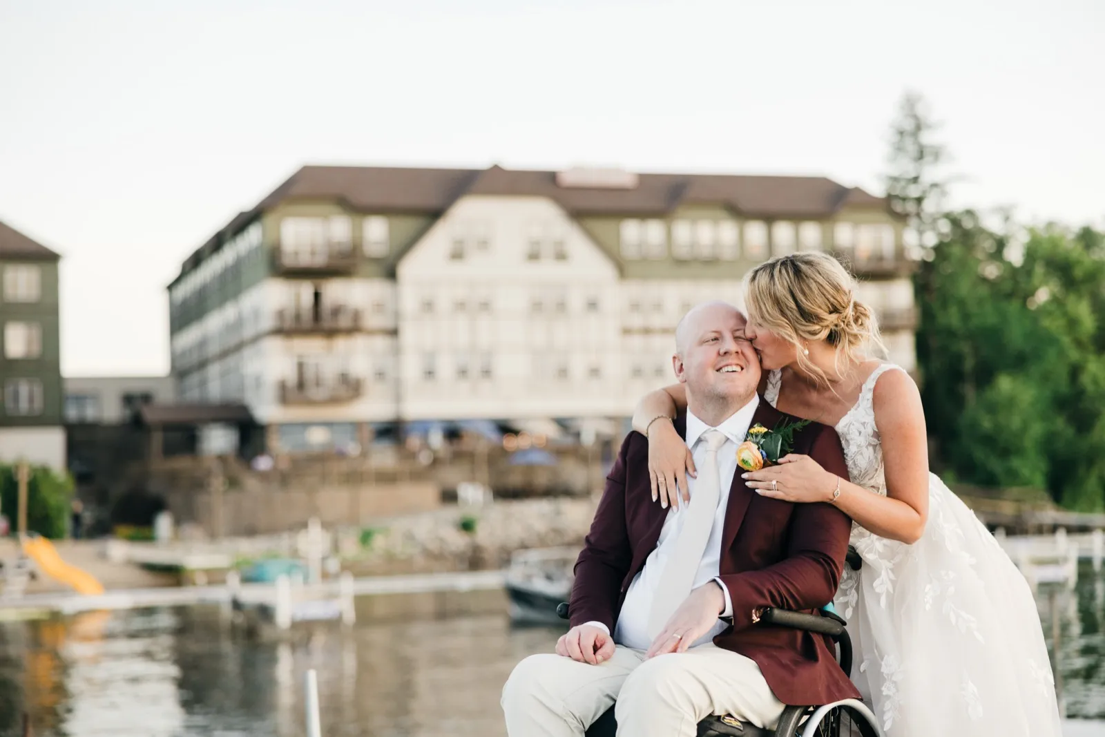 The bride leans down to kiss the groom's temple as he smiles broadly from his wheelchair on a dock, with the Chase on the Lake lodge softly blurred behind them in golden evening light. — Tim Larsen Photography, Brainerd Lakes MN