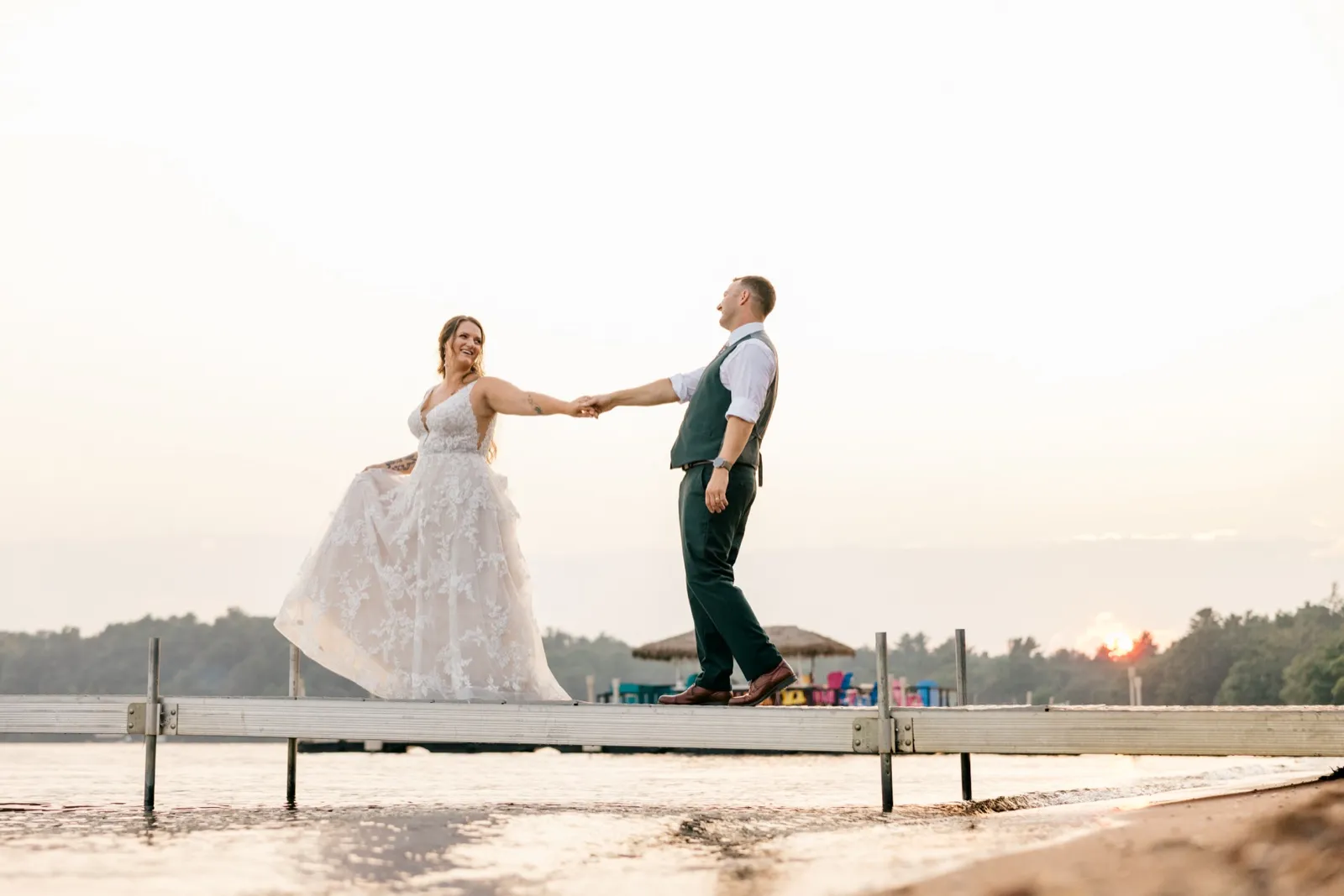 Chelsea and Eric hold hands while standing on a dock over Gull Lake at sunset, her gown billowing as he turns toward the light — Tim Larsen Photography, Brainerd Lakes MN