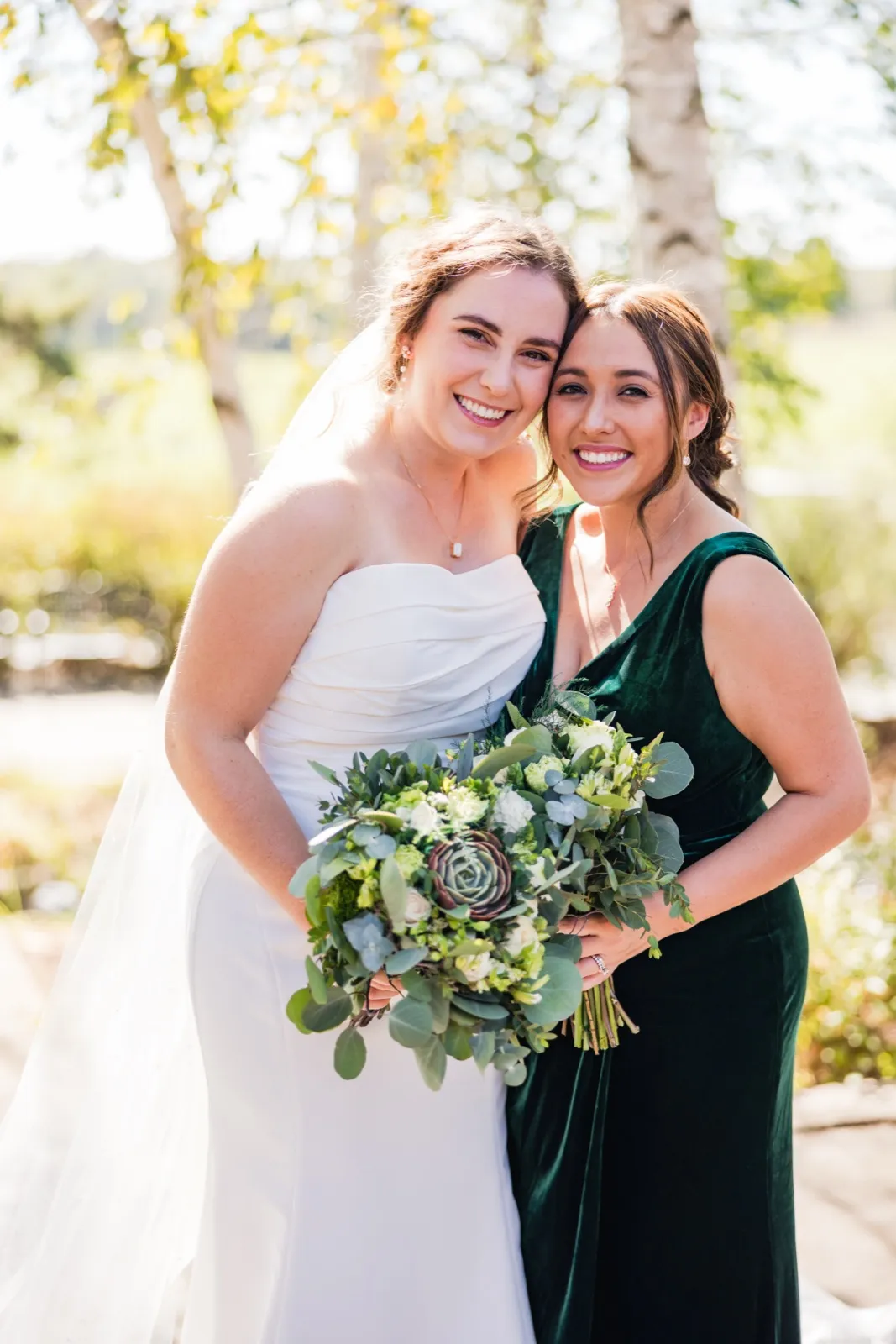 The bride and a bridesmaid in an emerald velvet dress lean cheek-to-cheek and smile at the camera, the bride holding a lush greenery-and-succulent bouquet under backlit trees — Tim Larsen Photography, Brainerd Lakes MN