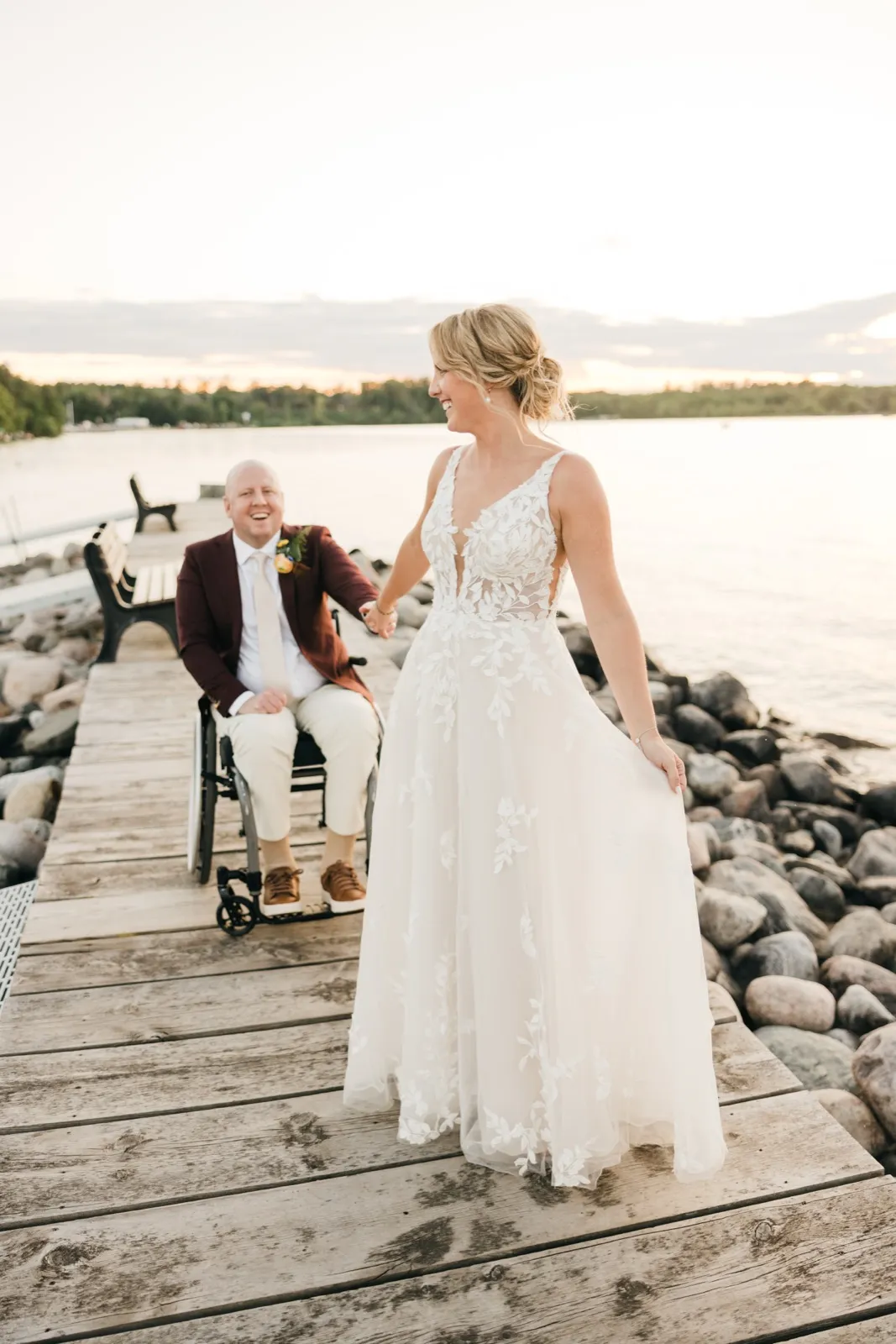 The bride in a lace gown leads her groom, who uses a wheelchair, down a wooden lakeside dock as he laughs, with calm water and trees behind them at sunset. — Tim Larsen Photography, Brainerd Lakes MN