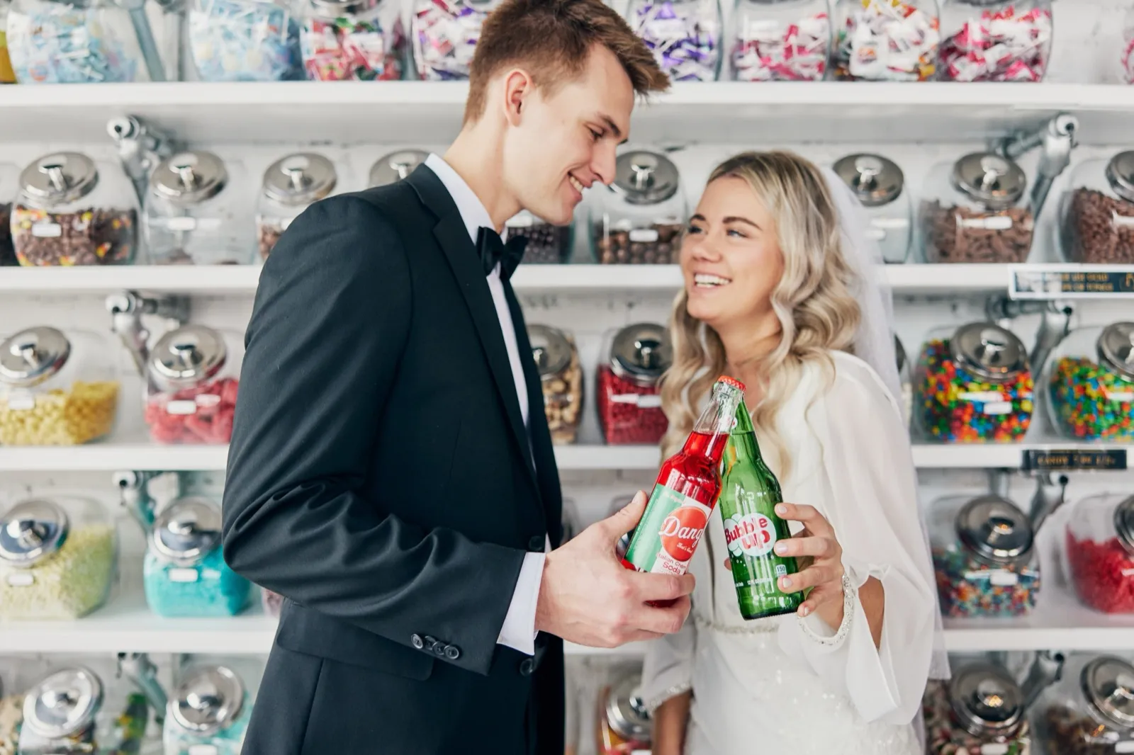 The bride and groom laugh together while holding vintage glass soda bottles in front of a wall of colorful candy jars. — Tim Larsen Photography, Brainerd Lakes MN