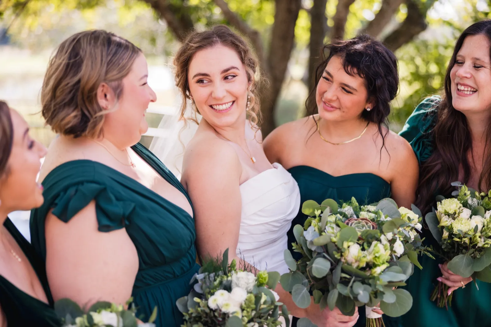 The bride laughs at the center of her bridesmaids in emerald dresses, all holding eucalyptus and white floral bouquets under dappled tree light — Tim Larsen Photography, Brainerd Lakes MN