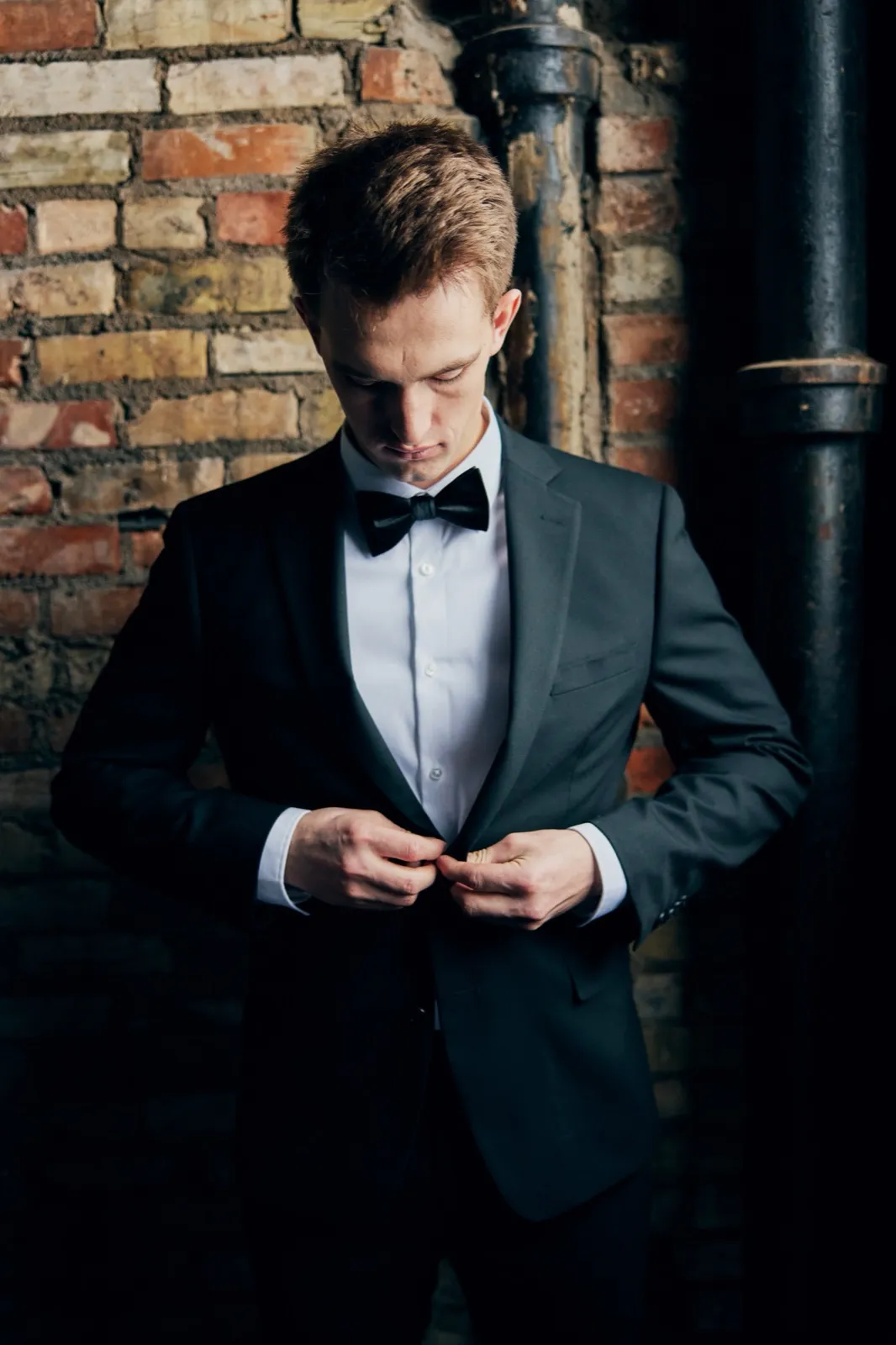 The groom in a black tuxedo and bow tie buttons his jacket while looking down, standing against an exposed brick wall with dark industrial piping. — Tim Larsen Photography, Brainerd Lakes MN