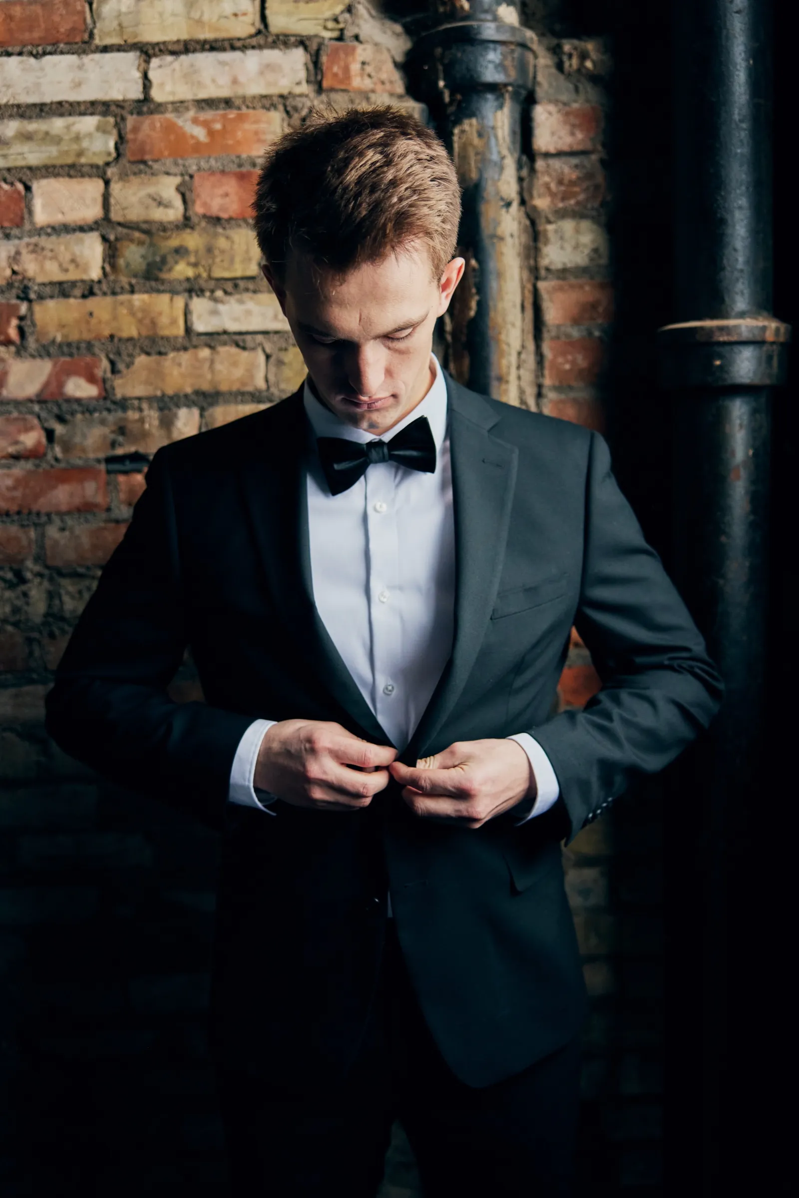 Sam in a black tuxedo and bow tie buttons his jacket against an exposed brick wall with dark industrial piping — Tim Larsen Photography, Brainerd Lakes MN