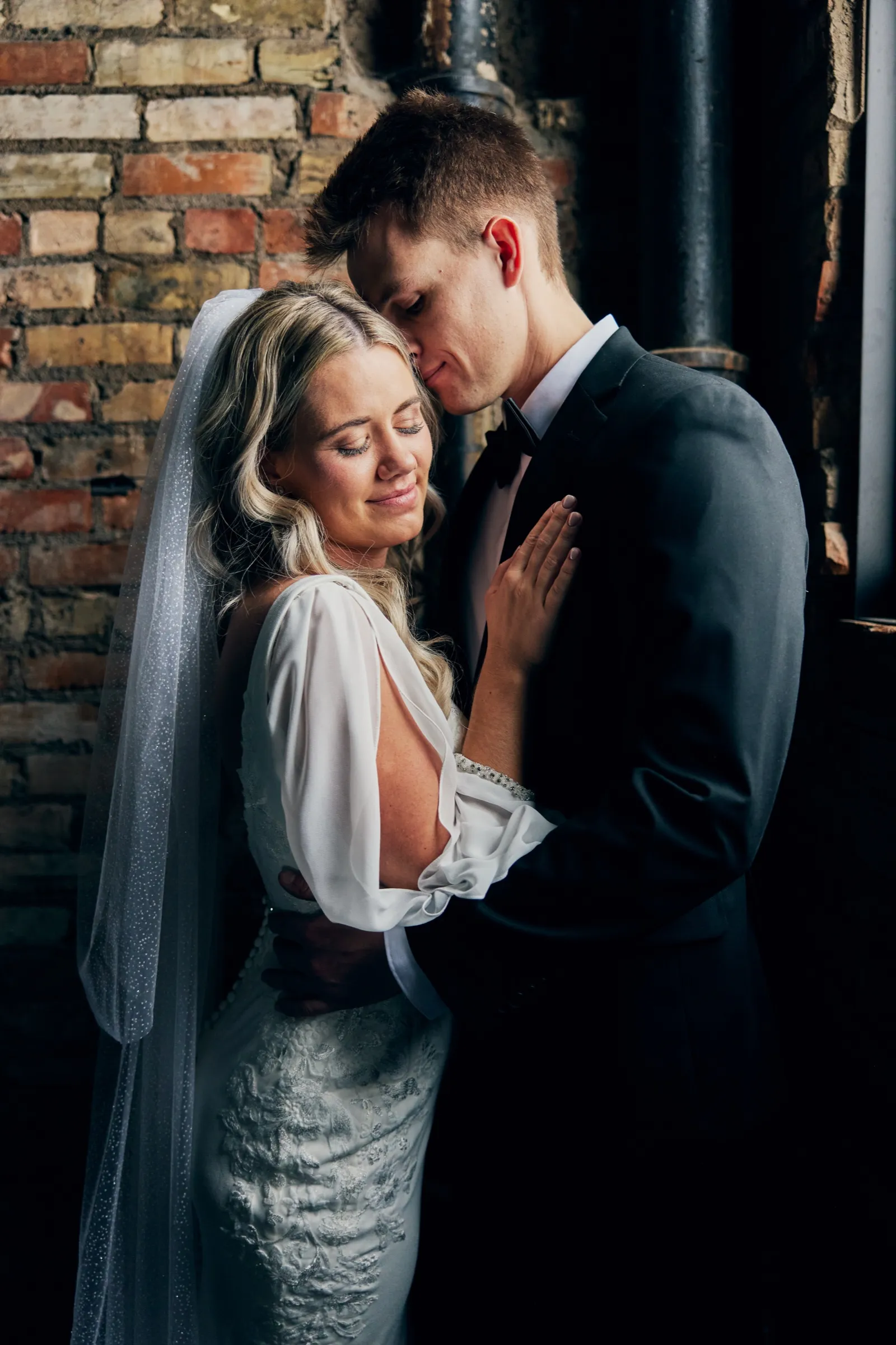 Madeline closes her eyes and smiles softly as Sam leans his face into her hair, the two embracing against a brick wall in window light — Tim Larsen Photography, Brainerd Lakes MN
