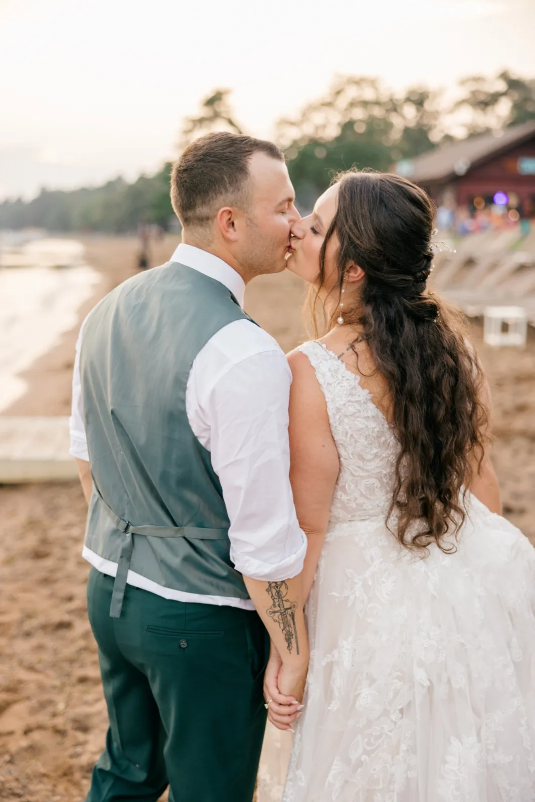 Chelsea and Eric kiss in profile on a sandy lakeshore at golden hour, with the lodge and bokeh string lights softly visible in the distance behind them — Tim Larsen Photography, Brainerd Lakes MN