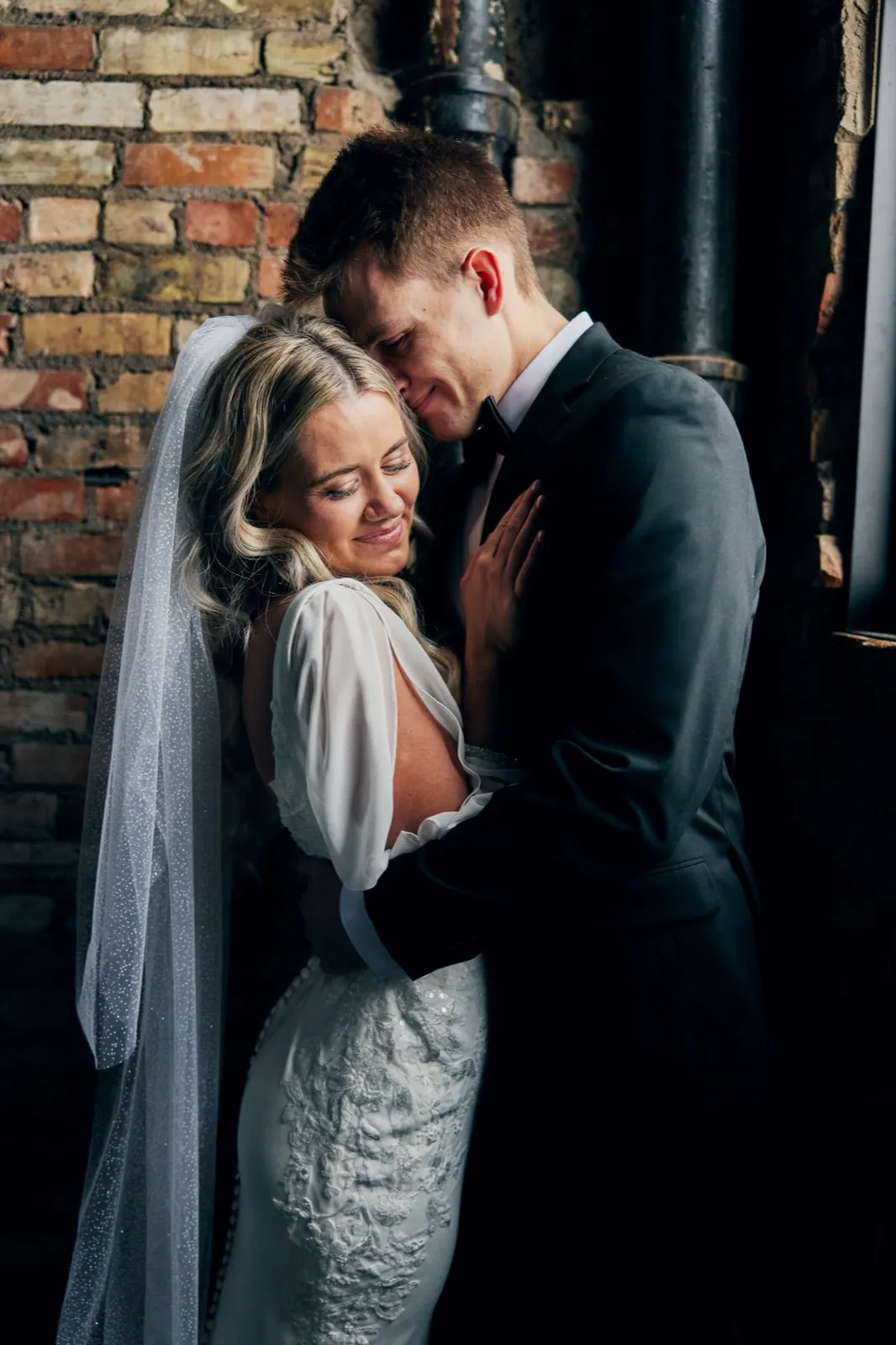 The bride rests her cheek against the groom's chest with eyes closed and a soft smile while he leans into her hair, both lit by window light against an exposed brick wall. — Tim Larsen Photography, Brainerd Lakes MN