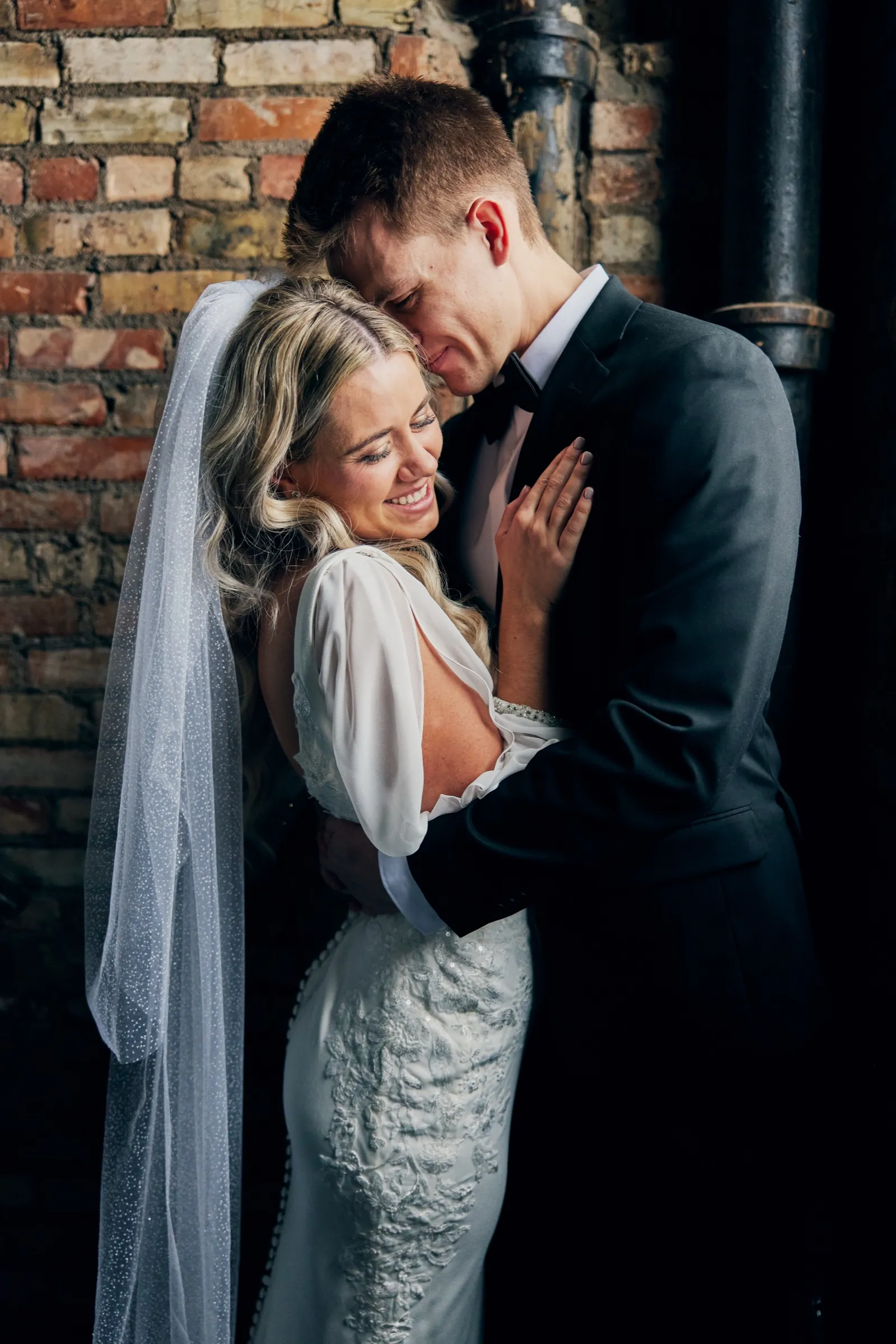 Madeline smiles with eyes closed as Sam leans his forehead to her hair, her veil falling beside them against an exposed brick wall — Tim Larsen Photography, Brainerd Lakes MN