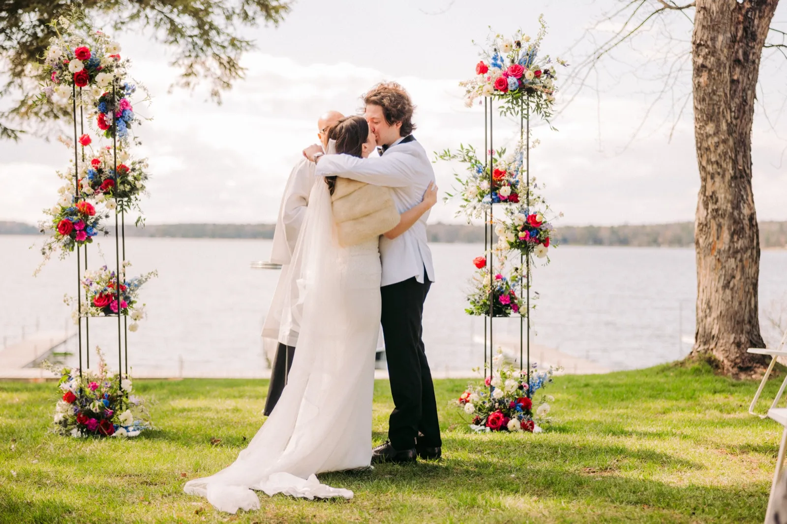 The bride and groom kiss between two tall pillar arrangements of bright florals while the officiant stands beside them, with a wide lake and bare-branched trees in the background — Tim Larsen Photography, Brainerd Lakes MN