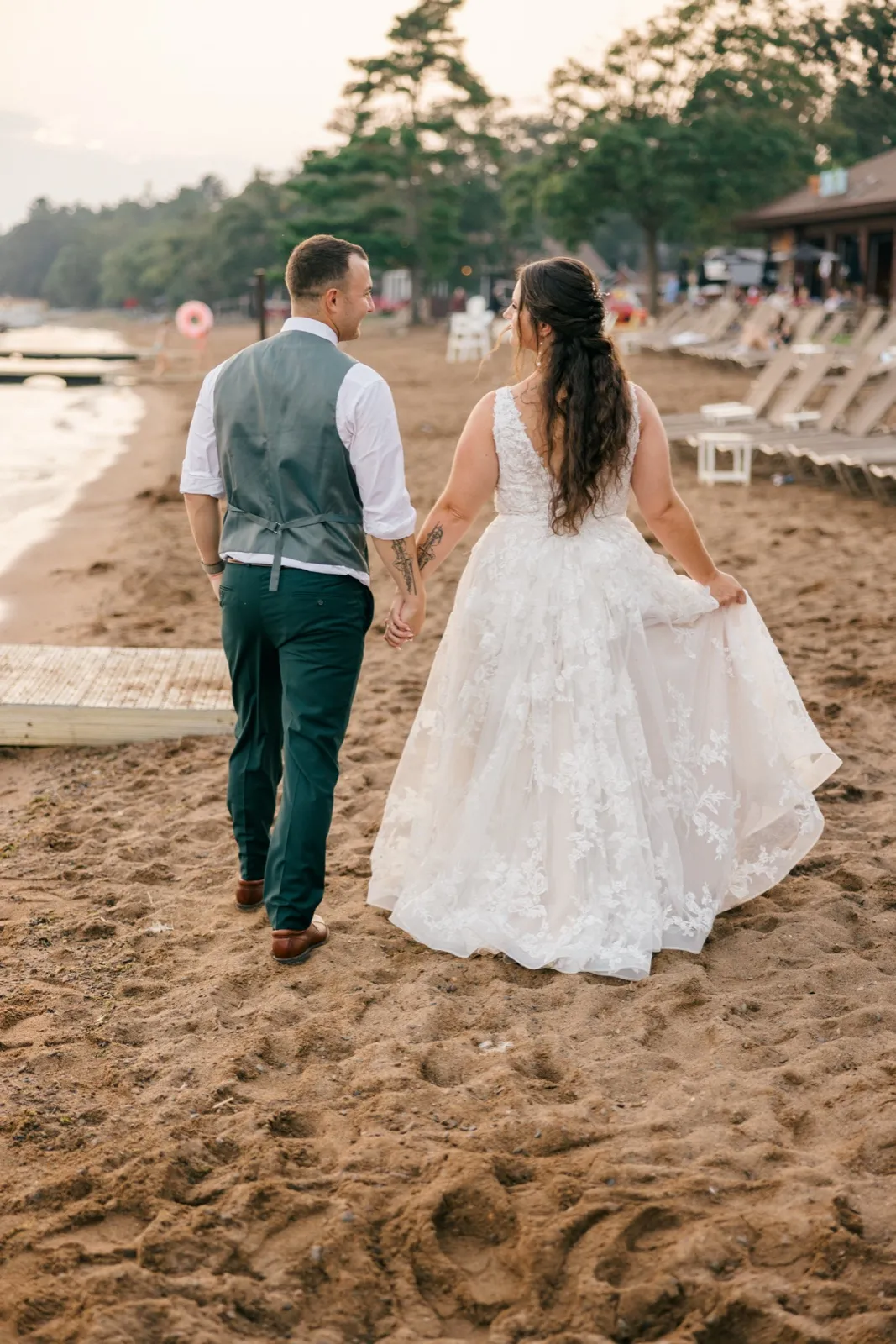 The bride glances back over her shoulder at the groom as they walk hand in hand across a sandy lakeshore beach, her train trailing in the sand