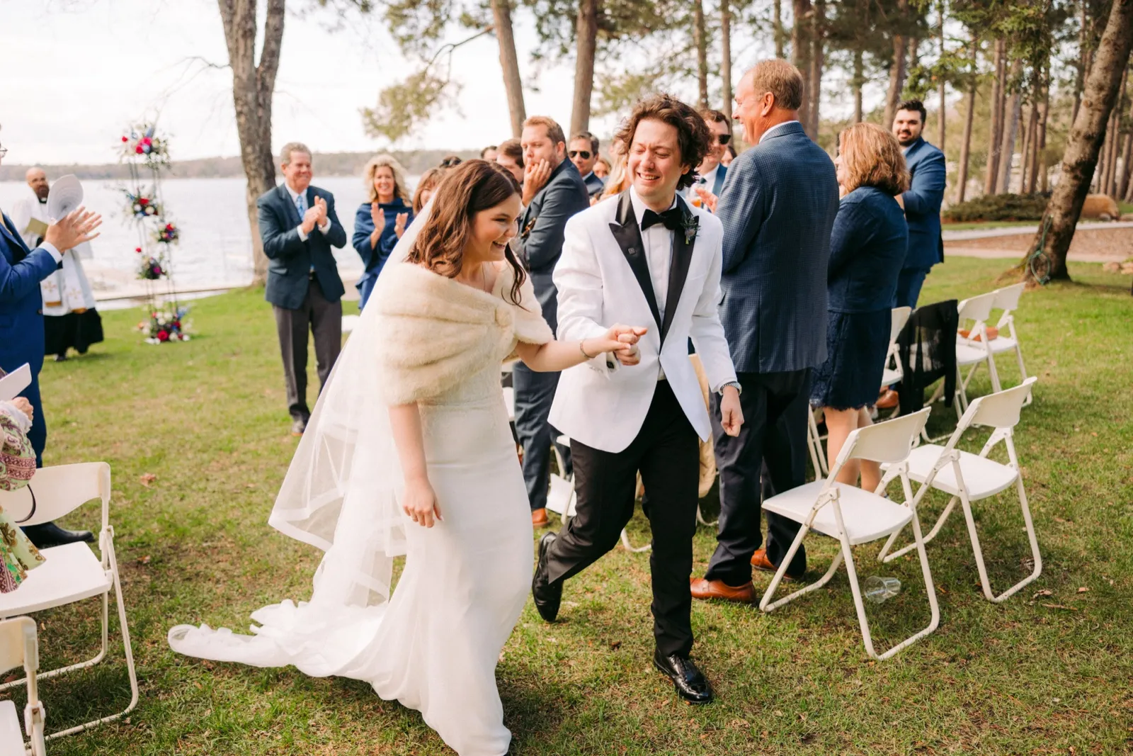 The bride and groom walk back down the aisle hand in hand, laughing, as guests applaud beside them at the lakeside ceremony — Tim Larsen Photography, Brainerd Lakes MN