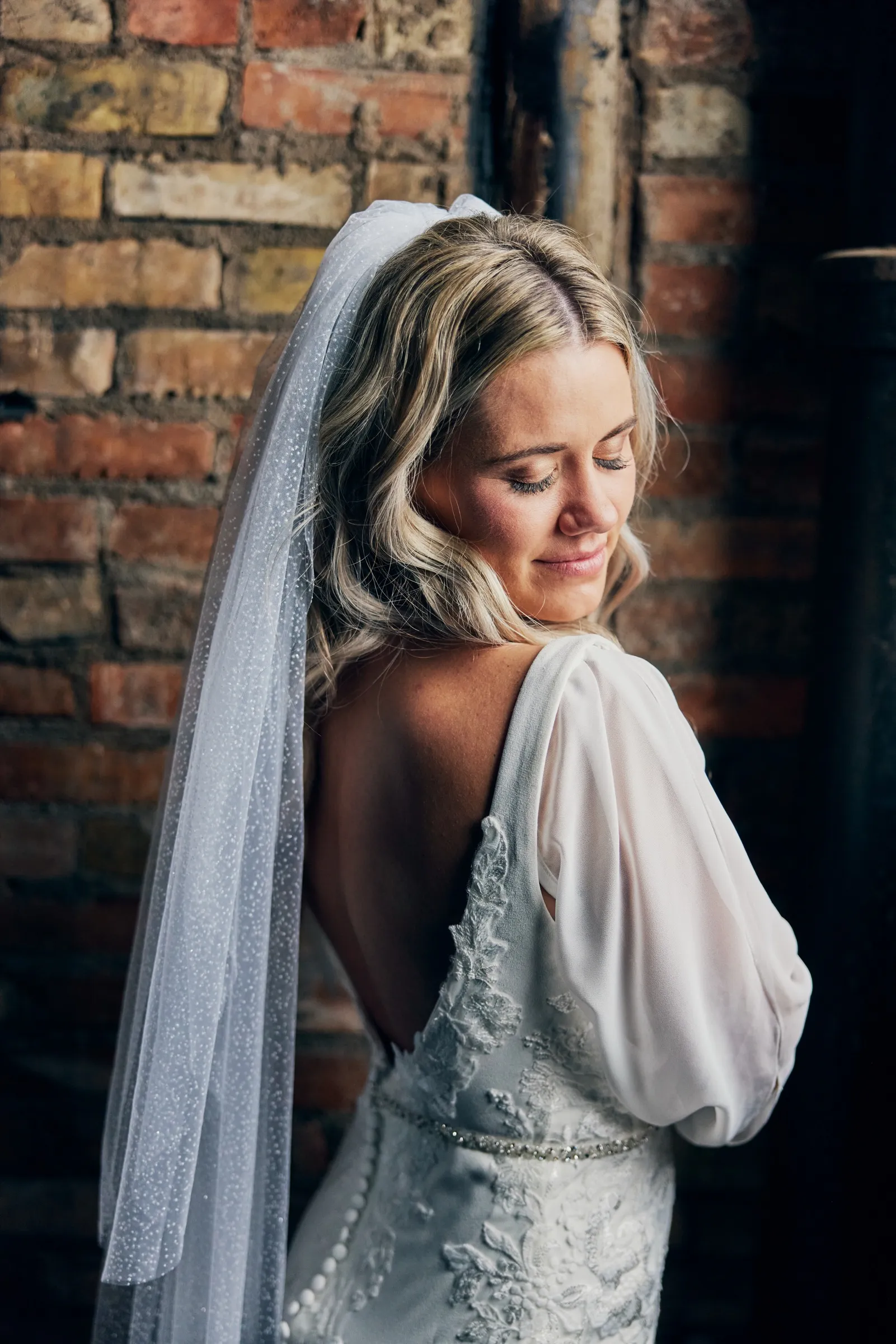 Madeline stands in profile, her sparkling veil and the lace back of her gown lit by window light against a warm exposed-brick wall — Tim Larsen Photography, Brainerd Lakes MN