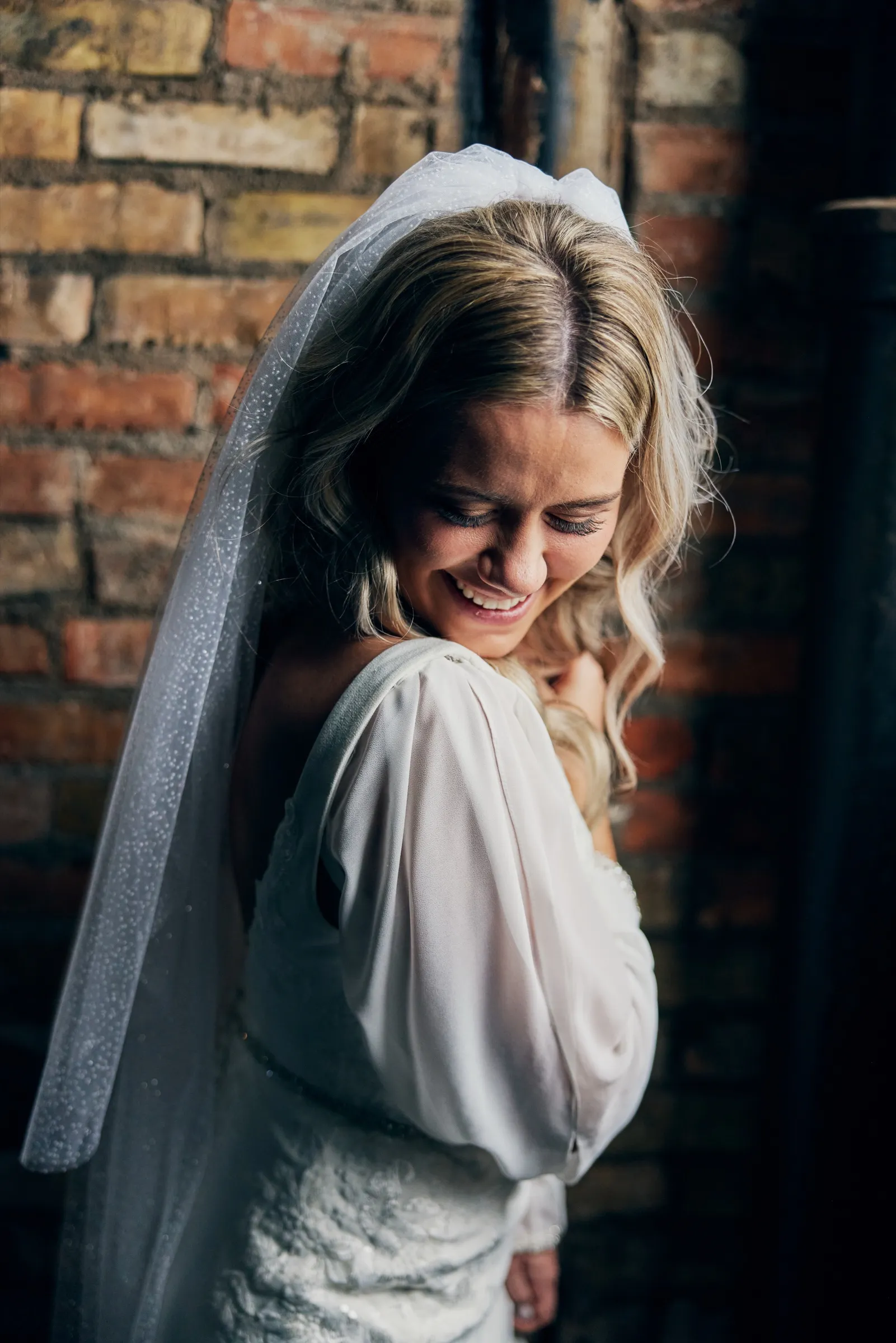 Madeline laughs with her head tilted down, her long veil falling past her shoulder against a textured brick wall — Tim Larsen Photography, Brainerd Lakes MN
