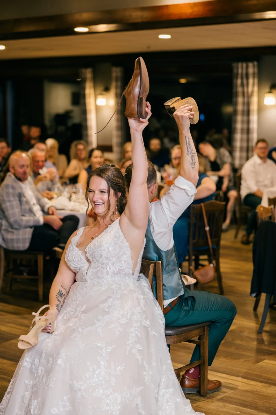 Chelsea laughs with her arms raised, holding her shoe in one hand and the groom's shoe in the other during the shoe game at the reception — Tim Larsen Photography, Brainerd Lakes MN