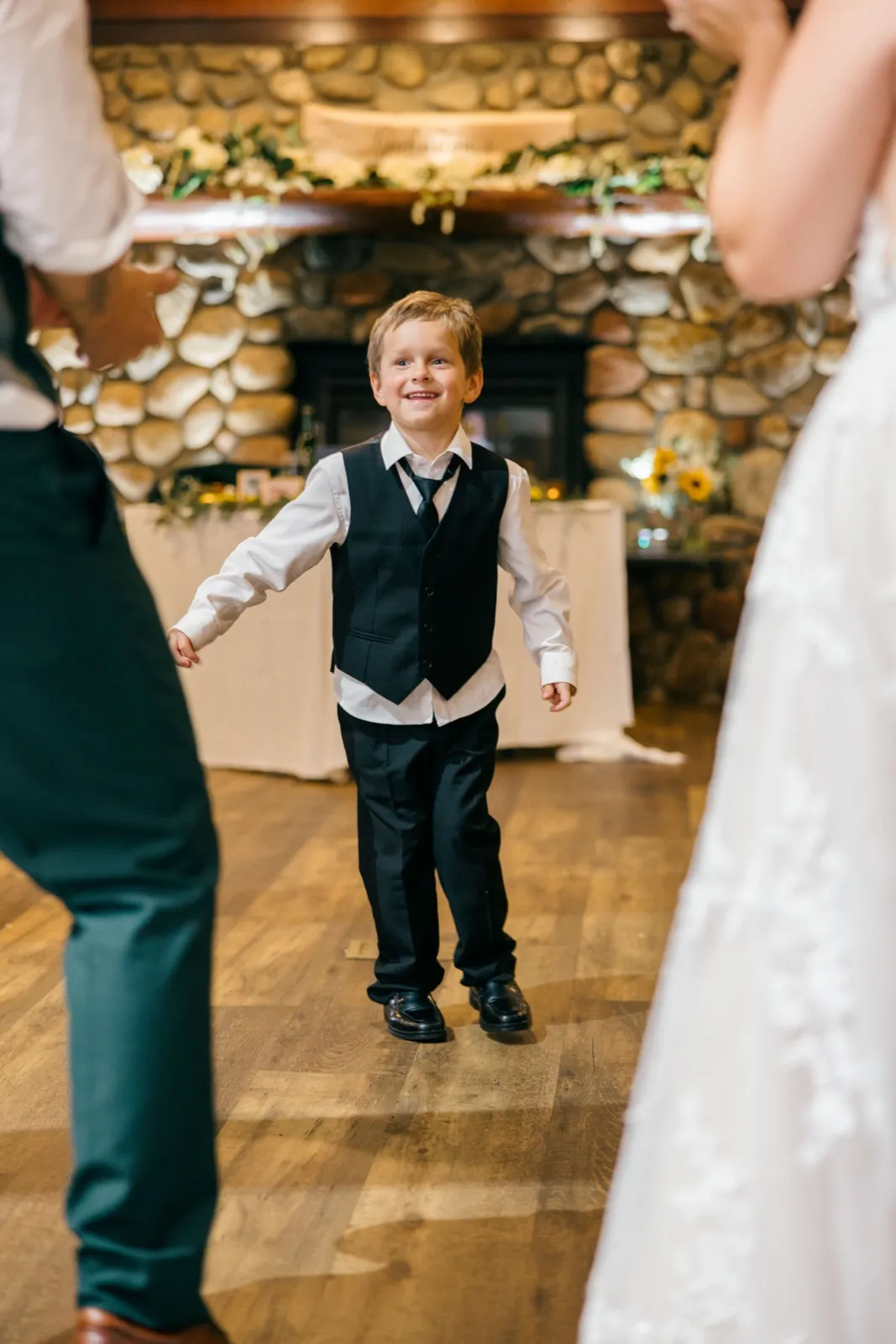 A young boy in a black vest and tie grins as he dances between the out-of-focus figures of Chelsea and Eric on the reception dance floor — Tim Larsen Photography, Brainerd Lakes MN