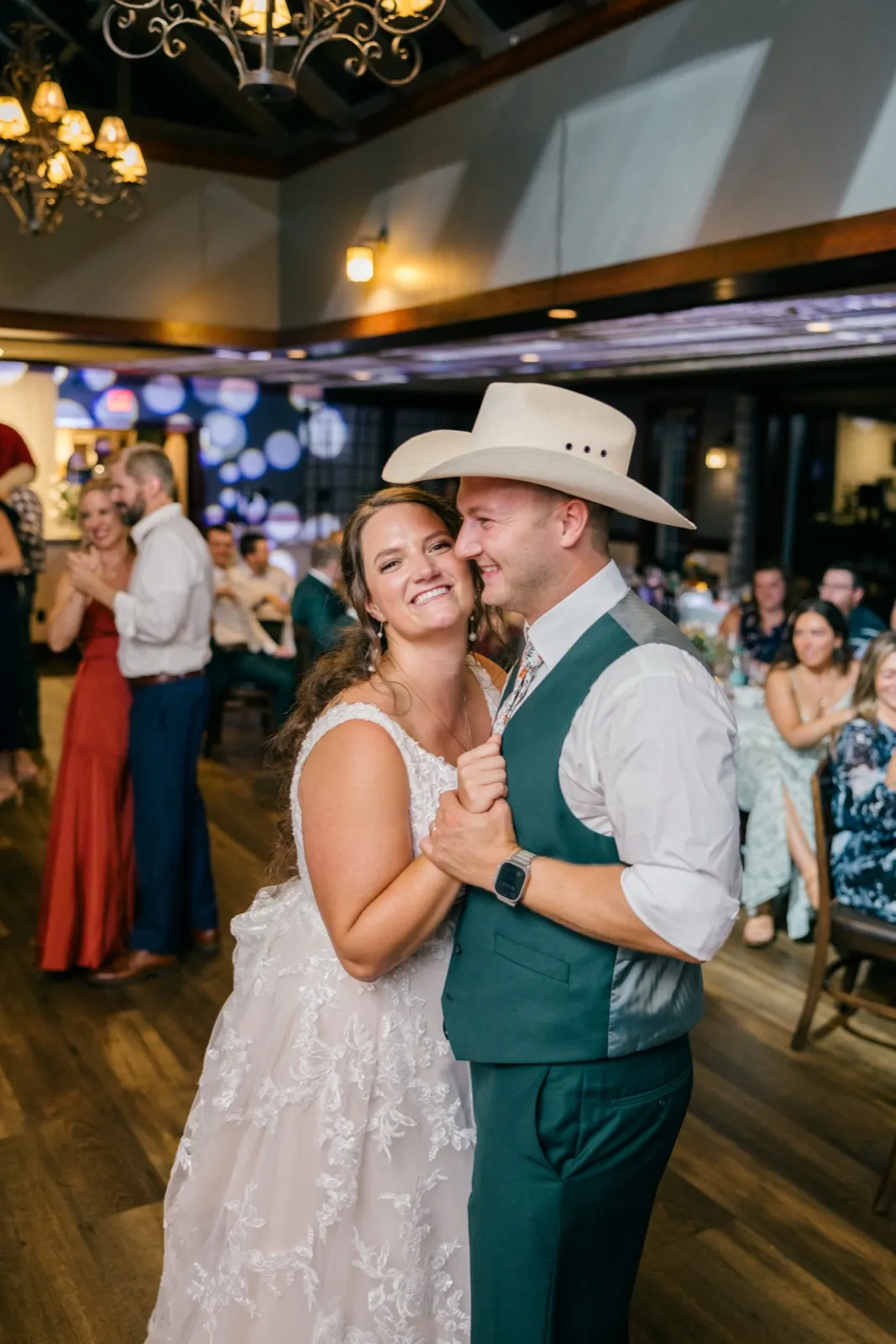Chelsea leans into Eric, who wears a cowboy hat and teal vest, both smiling broadly on the dance floor as guests look on beneath ornate iron chandeliers — Tim Larsen Photography, Brainerd Lakes MN