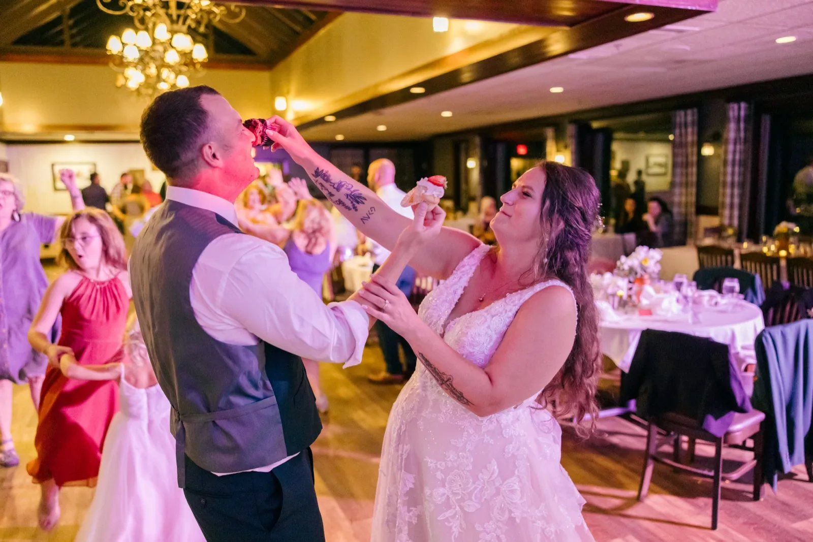 Chelsea and Eric simultaneously feed each other cake on the dance floor as guests dance around them under a chandelier at Grand View Lodge — Tim Larsen Photography, Brainerd Lakes MN