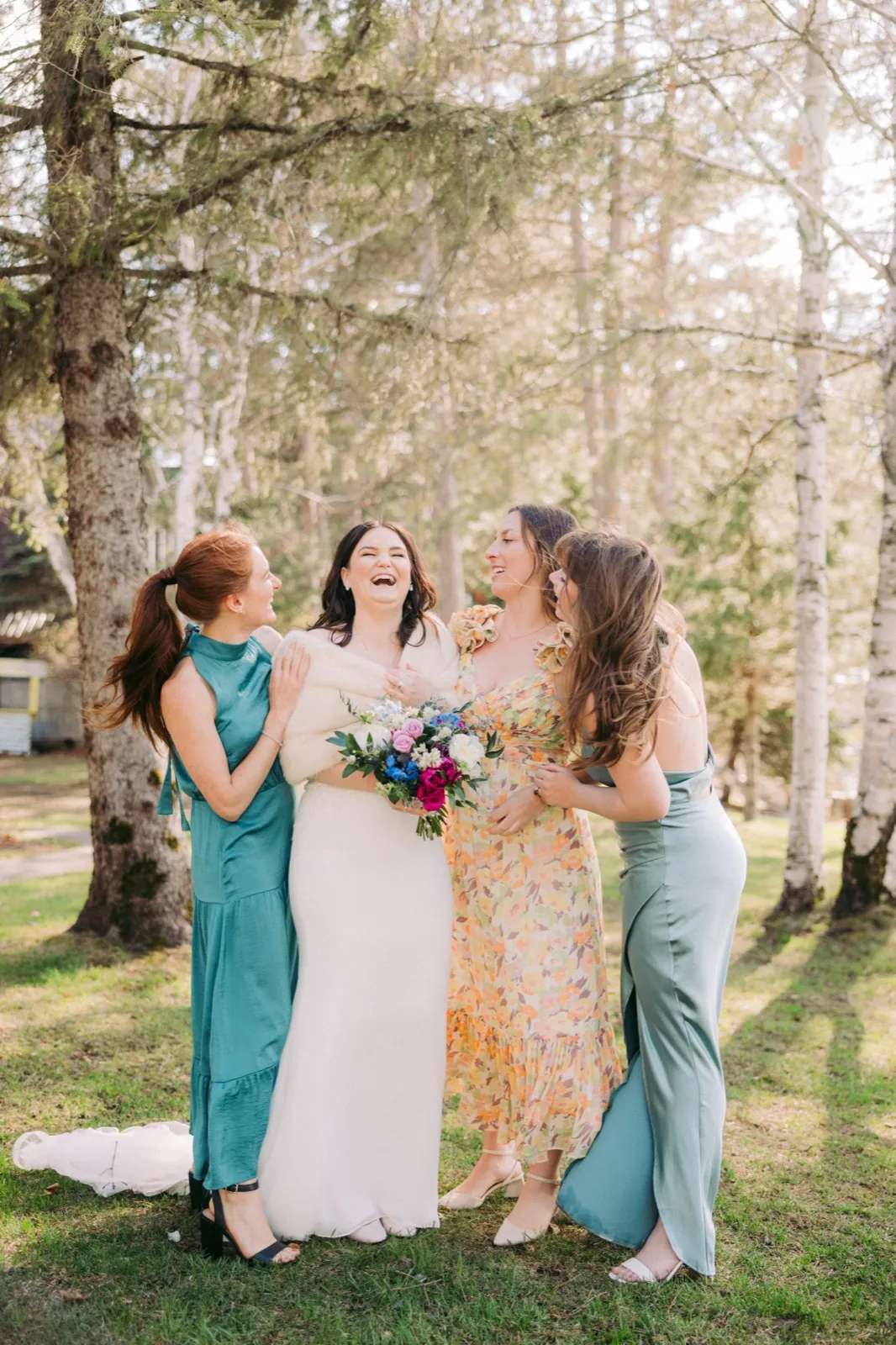 The bride laughs with her three bridesmaids in mismatched dresses among tall birch and pine trees, holding a colorful bouquet at Manhattan Beach Lodge — Tim Larsen Photography, Brainerd Lakes MN