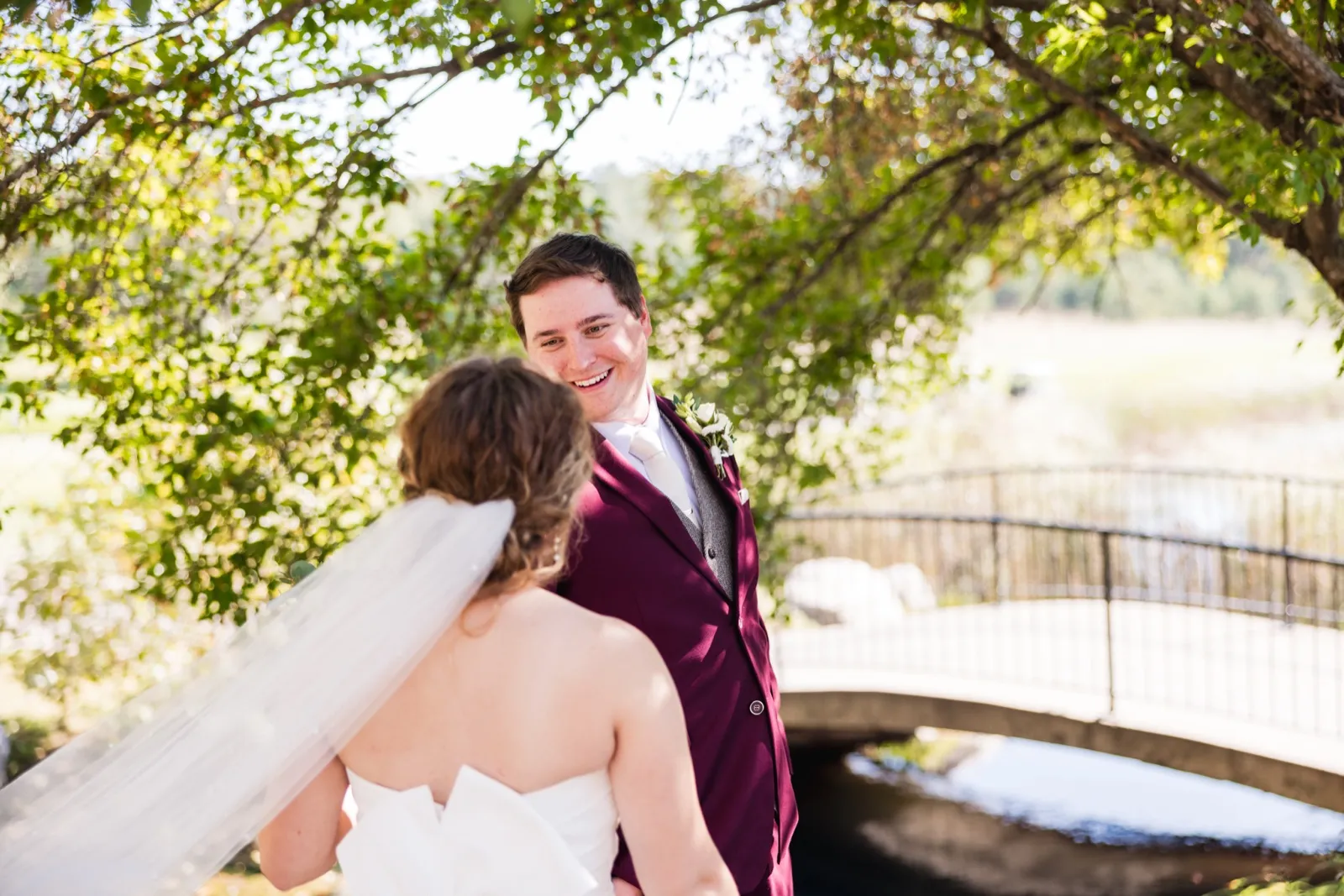 The groom in a burgundy suit smiles at his bride, seen from behind with her veil draped over her shoulder, on a garden bridge framed by overhanging leaves — Tim Larsen Photography, Brainerd Lakes MN