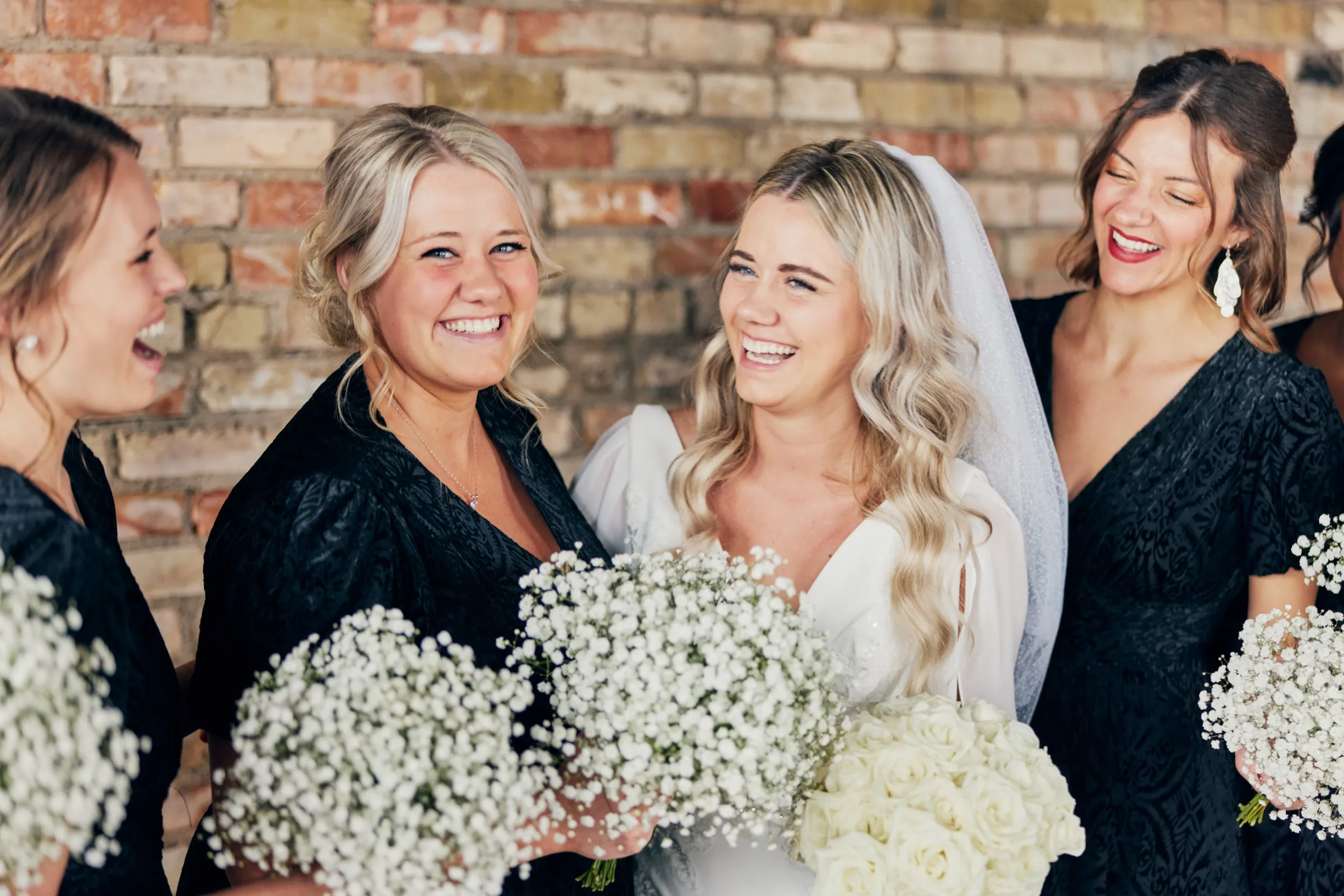 Madeline and her bridesmaids in black dresses laugh together in front of an exposed brick wall, holding white baby's breath and rose bouquets — Tim Larsen Photography, Brainerd Lakes MN