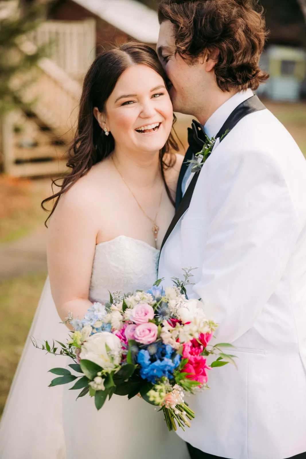 The bride laughs openly toward the camera while the groom kisses the side of her head, her colorful bouquet of pink, blue, and white blooms held in front of them — Tim Larsen Photography, Brainerd Lakes MN