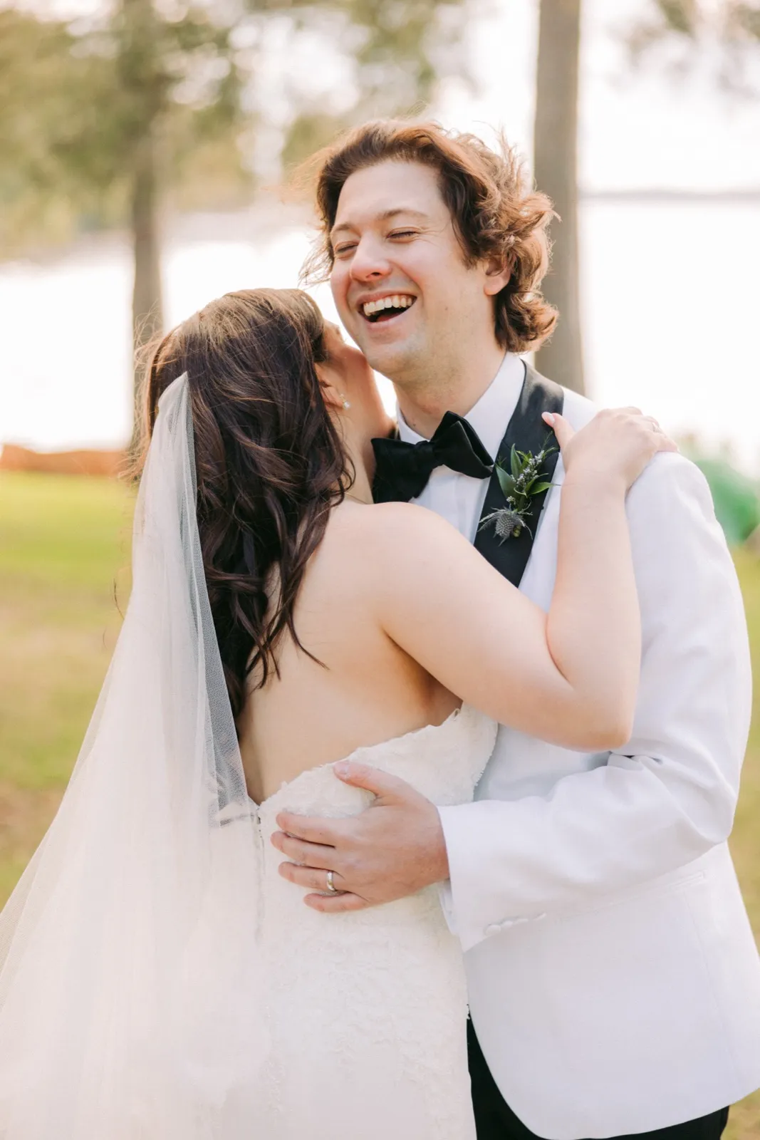 The groom laughs openly while the bride leans in close to whisper, her veil and hair lit from behind by afternoon sun on a lakeside lawn — Tim Larsen Photography, Brainerd Lakes MN