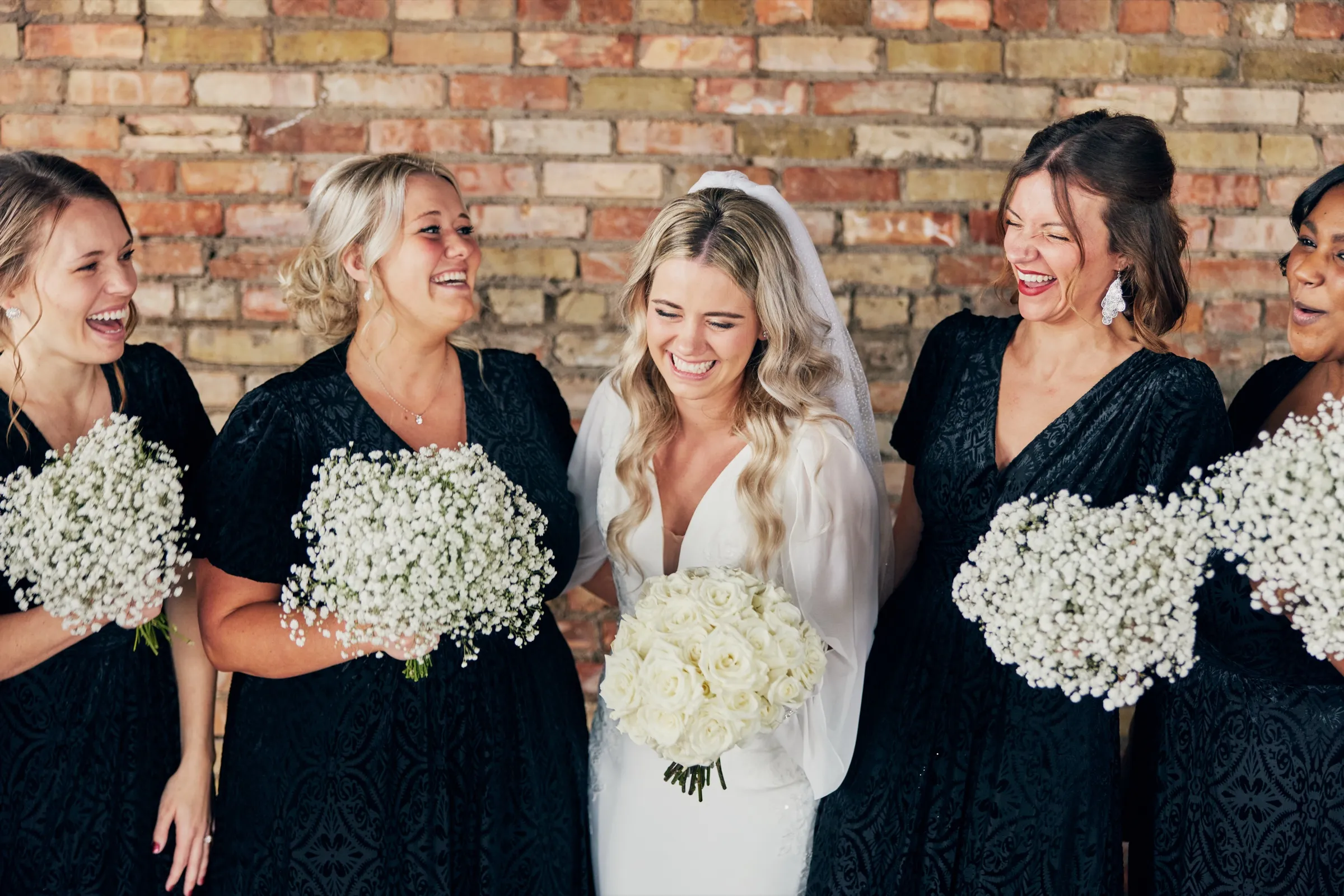 Madeline laughs with her eyes closed while her bridesmaids in black dresses flank her, all holding white bouquets against a brick wall — Tim Larsen Photography, Brainerd Lakes MN