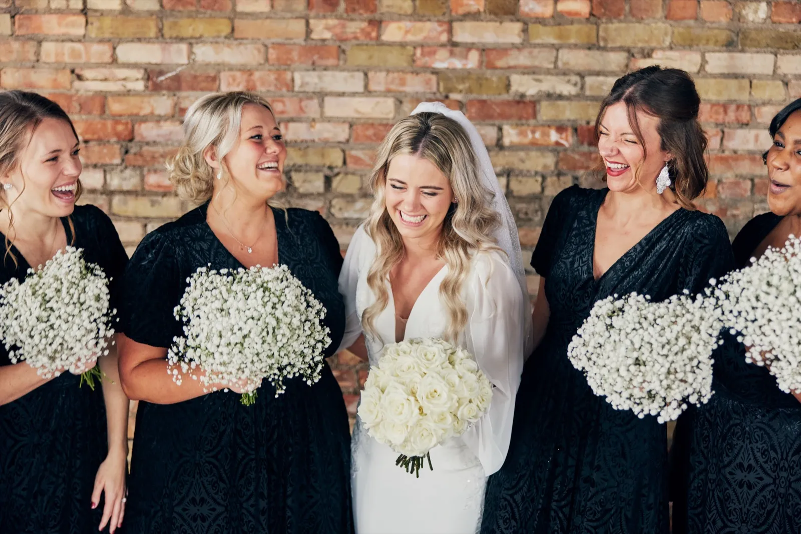 The bride laughs with her eyes closed while her bridesmaids in black dresses flank her, all holding white bouquets against an exposed brick wall. — Tim Larsen Photography, Brainerd Lakes MN