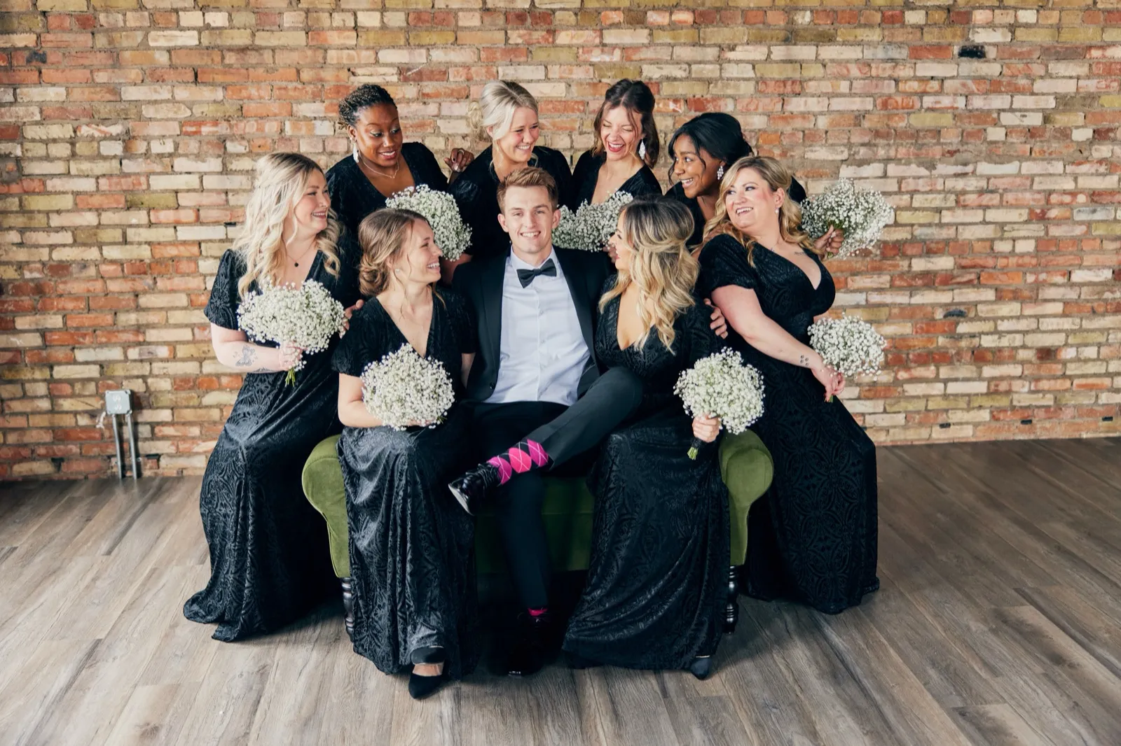 The groom sits on a green velvet sofa surrounded by seven bridesmaids in black dresses, all laughing together against an exposed brick wall. — Tim Larsen Photography, Brainerd Lakes MN