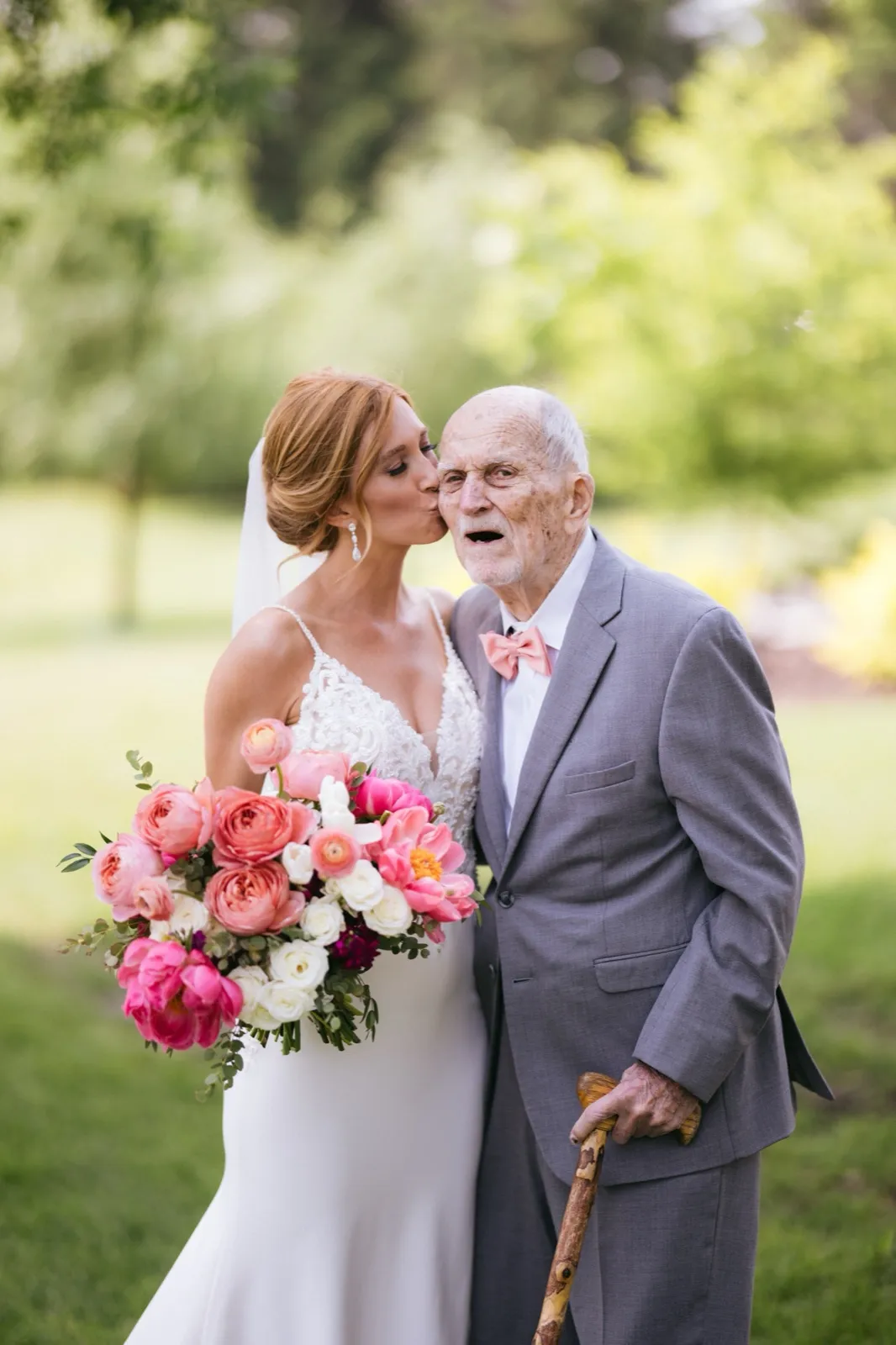 A bride kisses her elderly father on the cheek as he stands in a gray suit with a pink bow tie and a walking cane, holding her large pink and white bouquet on a green lawn — Tim Larsen Photography, Brainerd Lakes MN