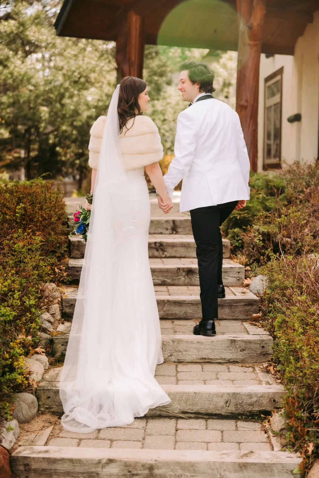 The bride and groom hold hands as they walk up stone steps toward the wooden archway entrance of Manhattan Beach Lodge, the bride's long veil trailing behind her — Tim Larsen Photography, Brainerd Lakes MN